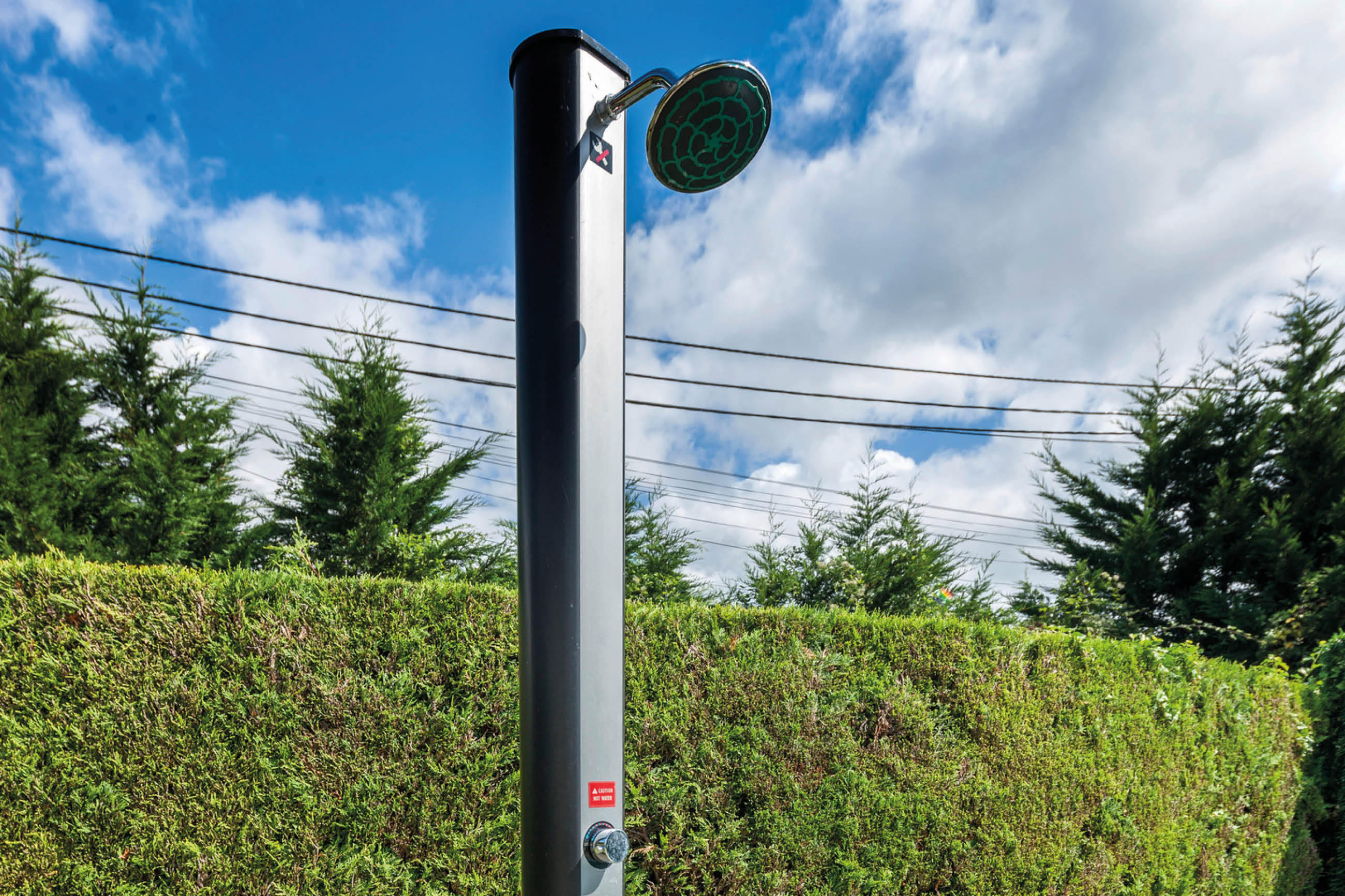 Schwarze Außendusche mit rundem Duschkopf,von GRE, vor grüner Hecke und blauem Himmel.