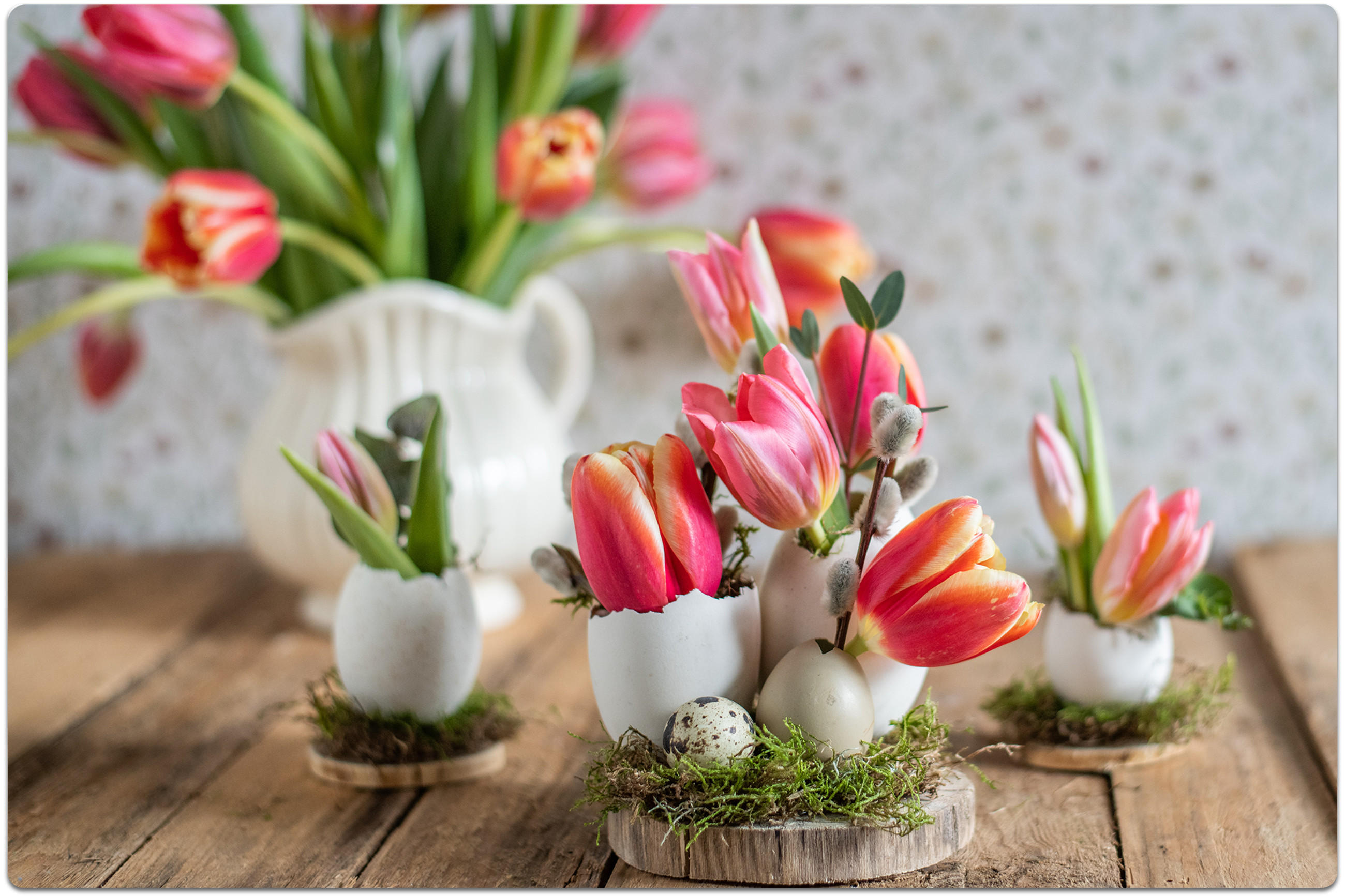 Frühlingsdeko auf Holztisch: Tulpen und Zweige in kleinen Eierschalen-Vasen auf Moos, im Hintergrund ein weißer Krug mit Tulpen.