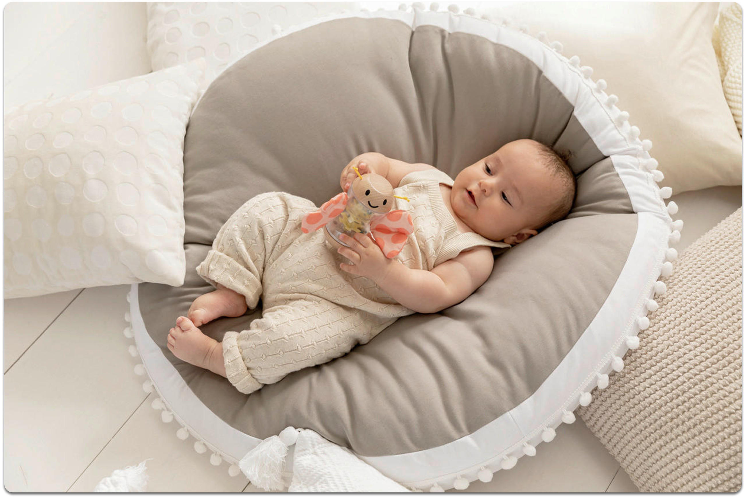 Ein Baby liegt auf einem beigen Babynestchen und hält dabei ein Spielzeug-Schmetterling aus Holz in der Hand. Das Baby trägt einen beigen Body mit Zopfmuster. Um das Babynest herum liegen weiße und beige Kissen.