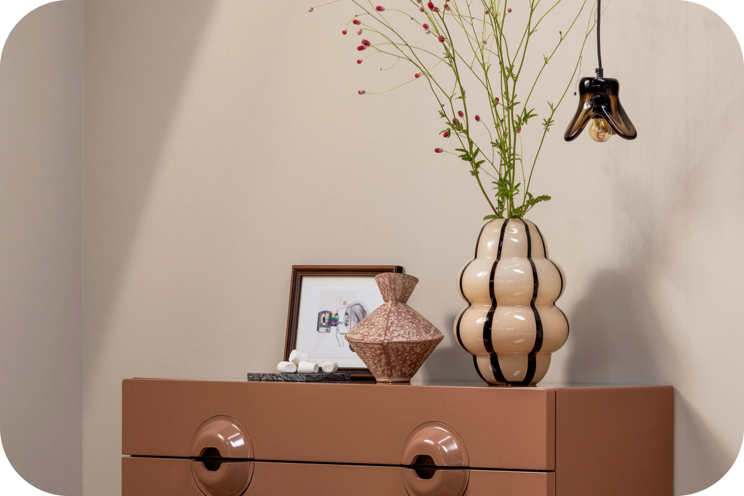 Lachsfarbenes Sideboard mit Vase, Bild und Deko; schwarze Hängelampe vor beiger Wand.