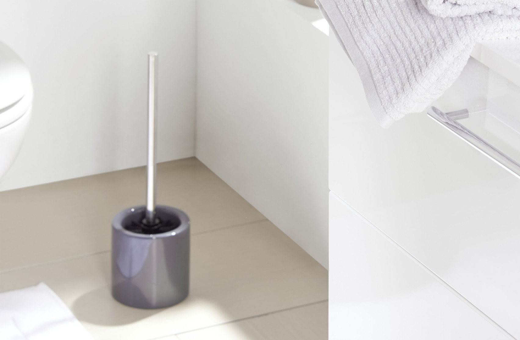 Šedý keramický stojan, držadlo na kefu, záchodová kefa, moderná kúpeľňa so svetlým obkladom a bielymi skrinkami, čistý a minimalistický interiér, biely uterák položený na umývadlovej skrinke