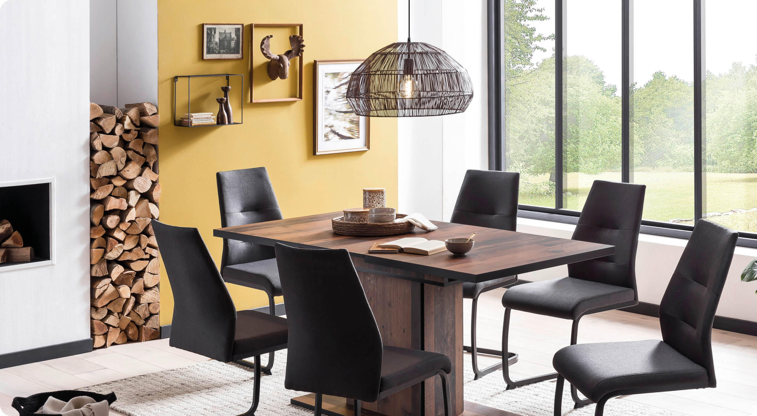 Esszimmer mit gelber Wand, Holztisch, sechs schwarzen Stühlen, Kamin mit Holzstapel und großen Fenstern mit Blick ins Grüne