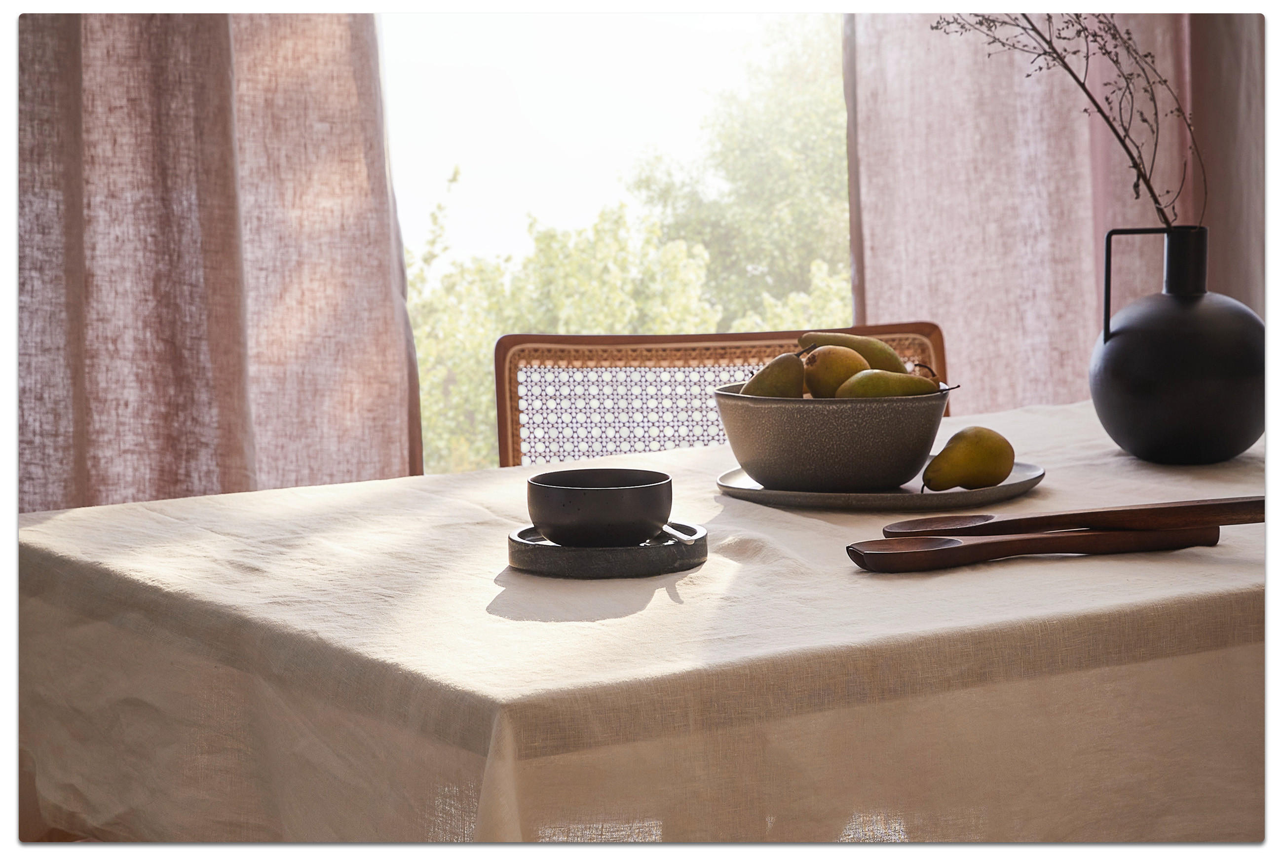 Esstisch mit Schale Birnen, schwarzer Tasse und Holzutensilien. Im Hintergrund ein Stuhl und lichtdurchlässige Vorhänge