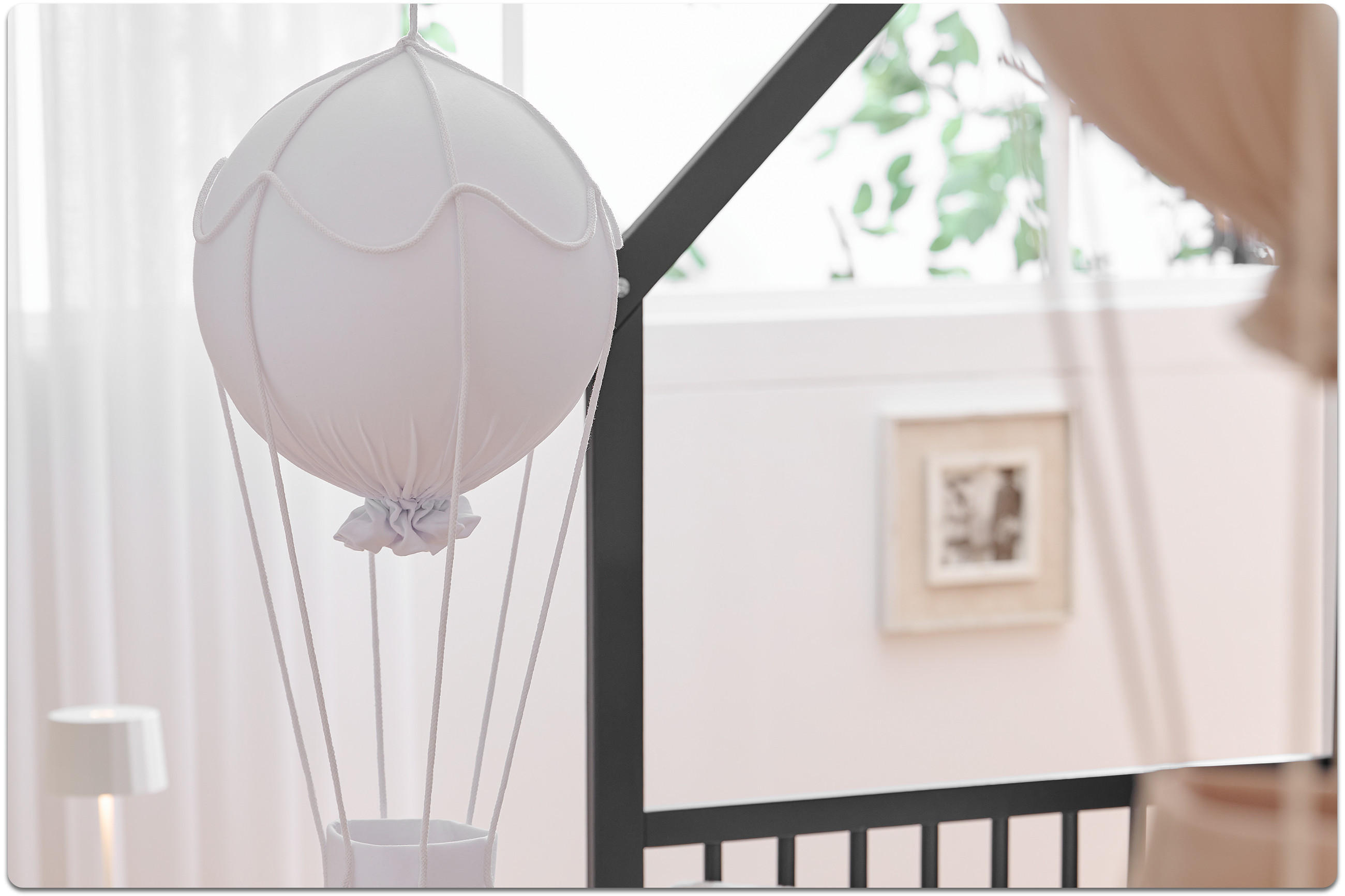 Dekoanhänger in weiß in Form eines Heißluftballons hängt vor einem Babybett mit schwarzem Bettgestell. Im Hintergrund sind weiße Vorhänge, eine Nachttischlampe und ein kleines, quadratisches Bild.