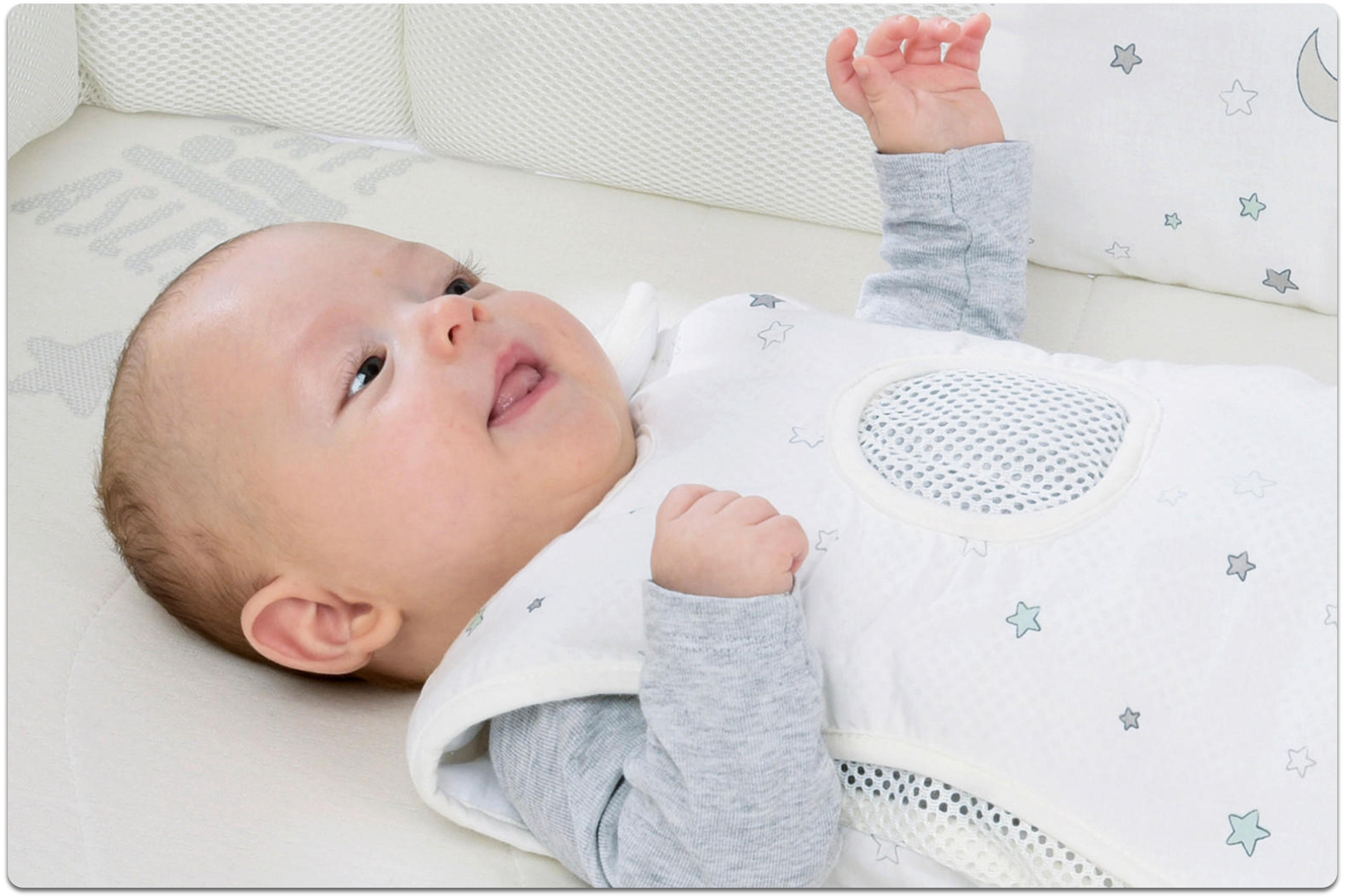 Baby liegt im Bett auf heller Matratze und trägt einen weißen Schlafsack mit Sternmuster und atmungsaktivem Mesh-Einsatz, dazu graues Langarmshirt.