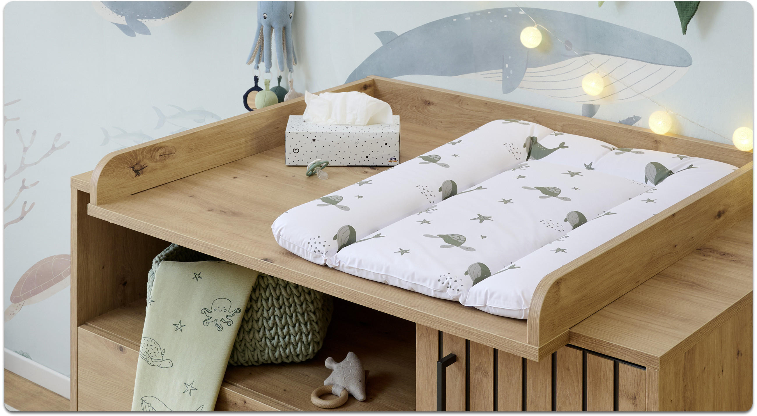 Holz-Wickelaufsatz mit weißer Wickelauflage mit Wal- und Pflanzenmotiven, daneben Taschentuchbox; darunter offene Fächer mit Decke, Korb und Spielzeug vor Wand mit Walillustration und Lichterkette.