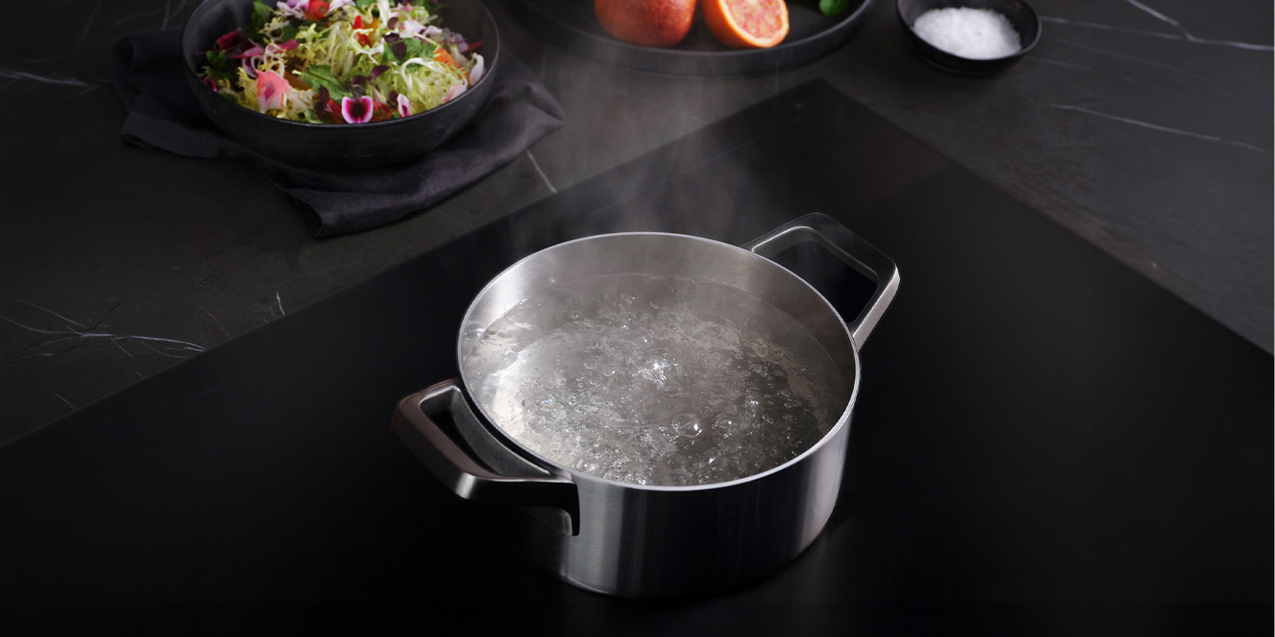 Kochtopf mit sprudelnd kochendem Wasser auf einem mattschwarzen AEG Induktionskochfeld; auf der dunklen Arbeitsfläche stehen Schale mit Salat, Grapefruit und Salz, während aufsteigender Dampf sichtbar ist.