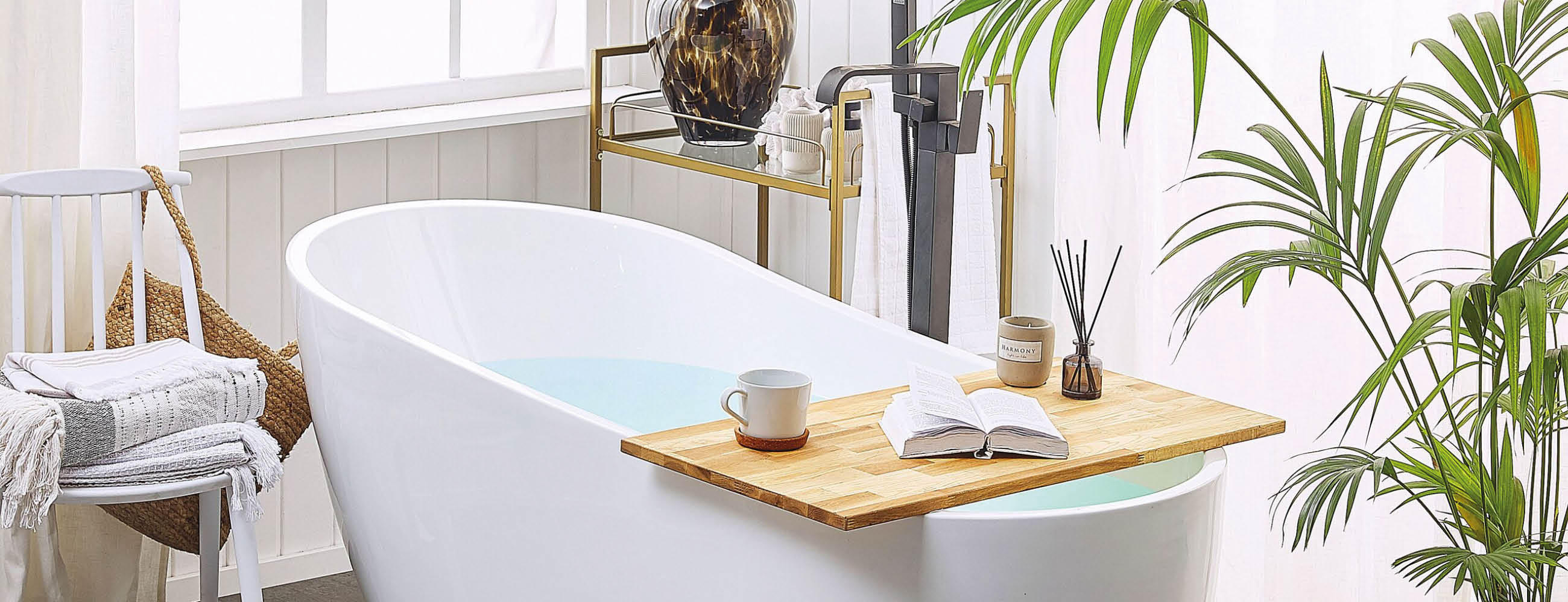 Freistehende weiße Badewanne mit Holzablage, auf der eine Tasse, ein offenes Buch und Duftstäbchen stehen, umgeben von Pflanzen und einem Handtuch auf einem Stuhl