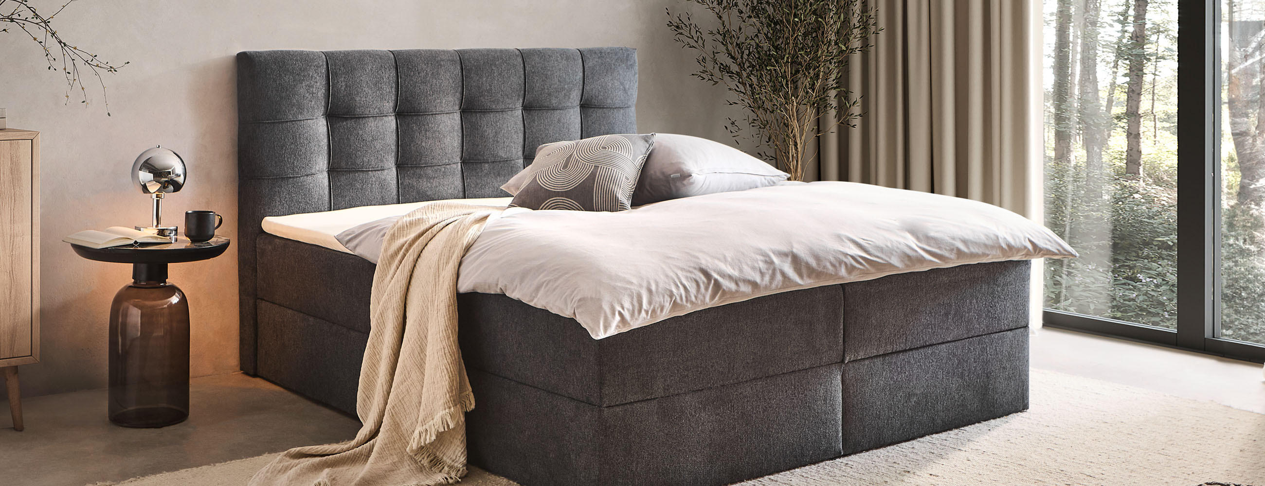 Dunkelgraues Boxspringbett mit Schlaftextilien, Nachttisch und Lampe gemütlich eingerichtet