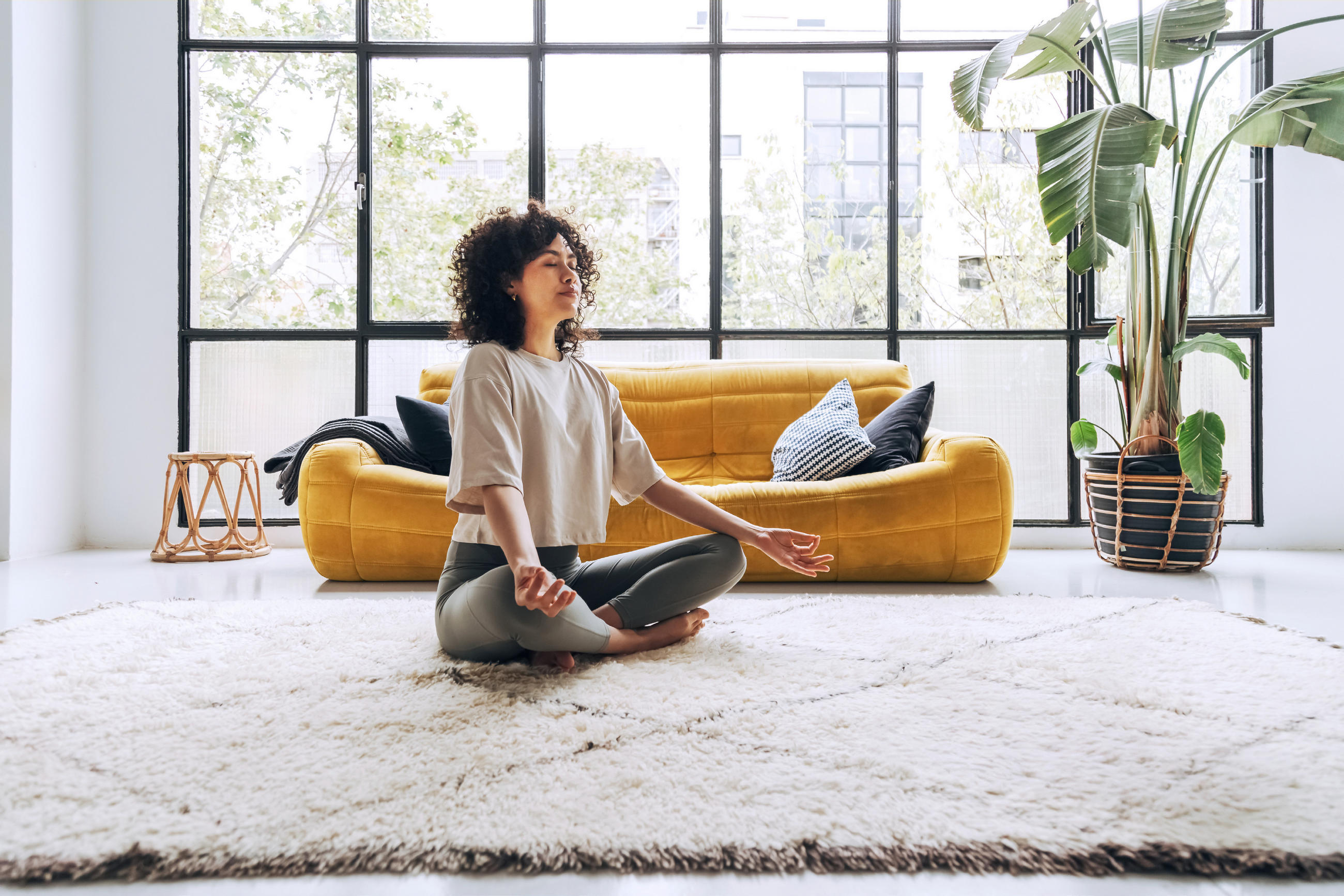 Eine Person sitzt meditierend auf einem hellen, weichen Teppich in einem modernen Wohnraum mit großer Fensterfront und gelbem Sofa.