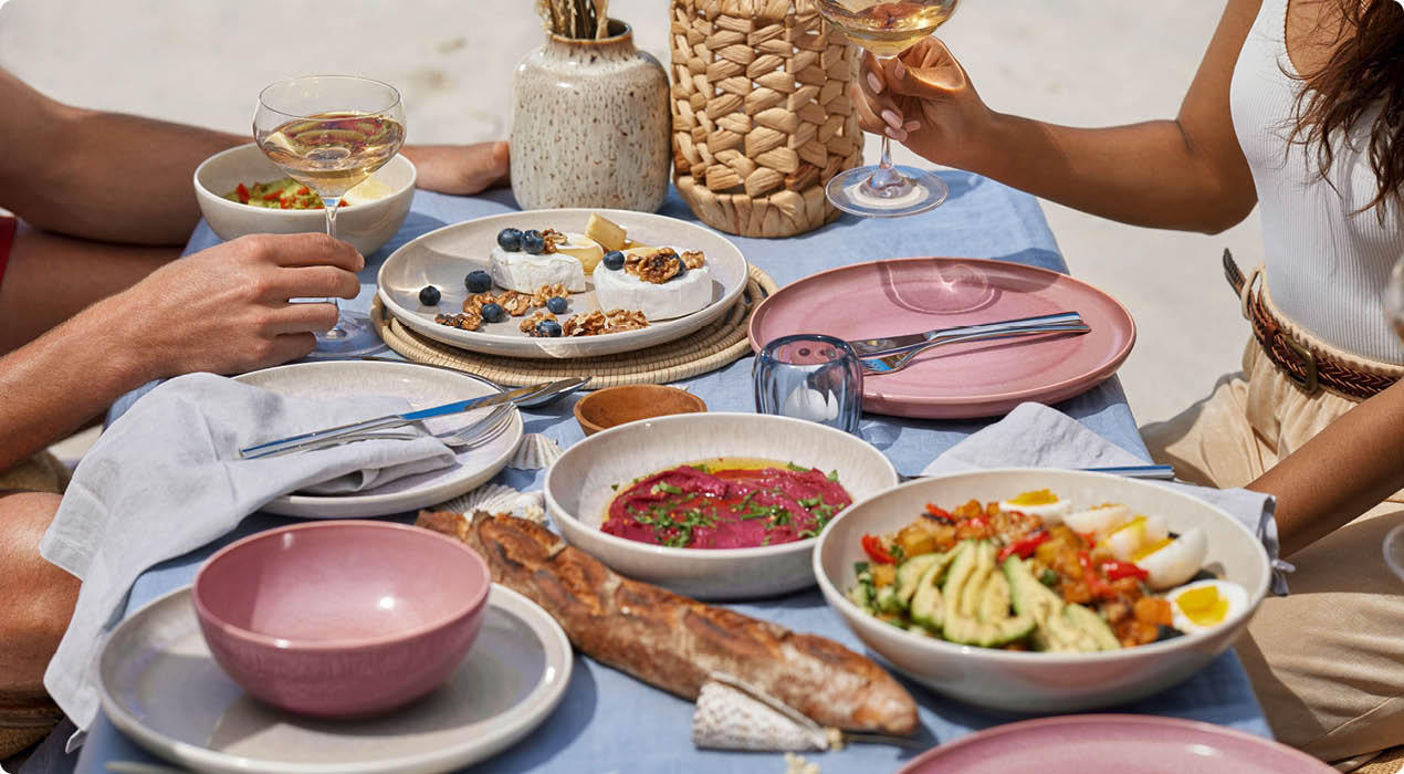 Esstisch mit blauer Tischdecke, gedeckt mit rosa und weißen Tellern, Schalen mit verschiedenen Speisen, Baguette, Weingläsern und zwei Personen, die anstoßen.