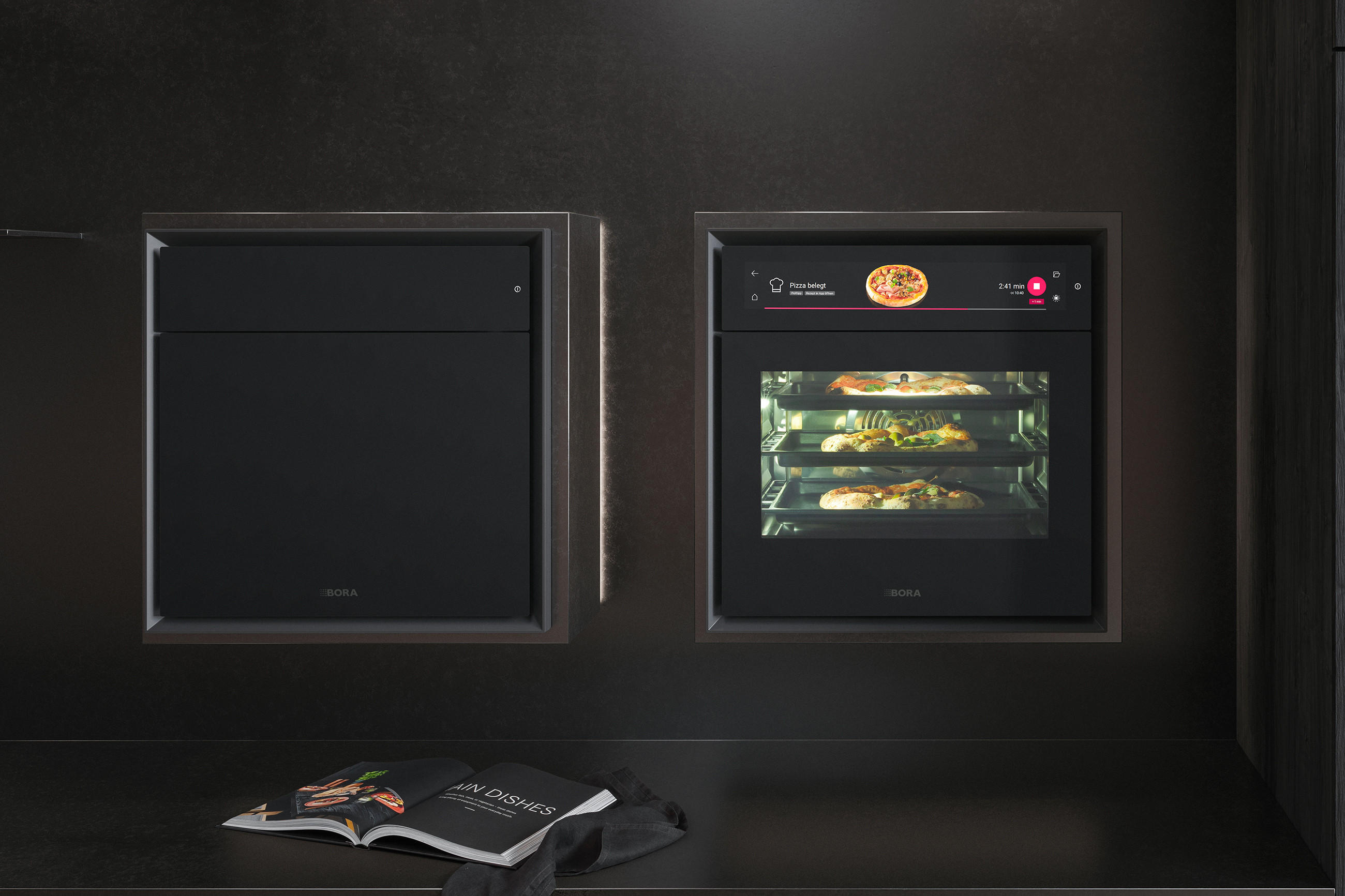 Zwei schwarze BORA Einbaugeräte in dunkler Küchenwand; rechts ein Backofen mit Display, in dem mehrere Pizzen gleichzeitig auf drei Ebenen gebacken werden.