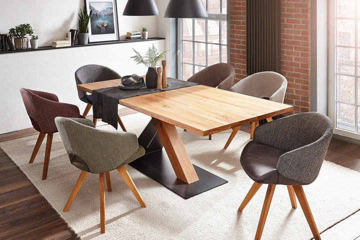 Stôl drevený, stoličky čalúnené rôznofarebné, závesné lampy čierne, kvetináče sivé, dekorácie zelené, obraz na stene, okno veľké, koberec svetlý, stena biela, podlaha drevená.