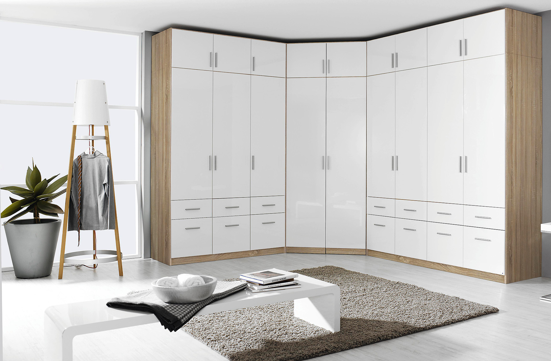 rohová skriňa, biela lesklá farba, drevený detail, biele skrine, úložný priestor, zásuvky, dvierka, lampa, vešiak, sivá košeľa, kravata, svetlohnedý koberec, nízky biely konferenčný stolík, knihy, zelená rastlina v kvetináči