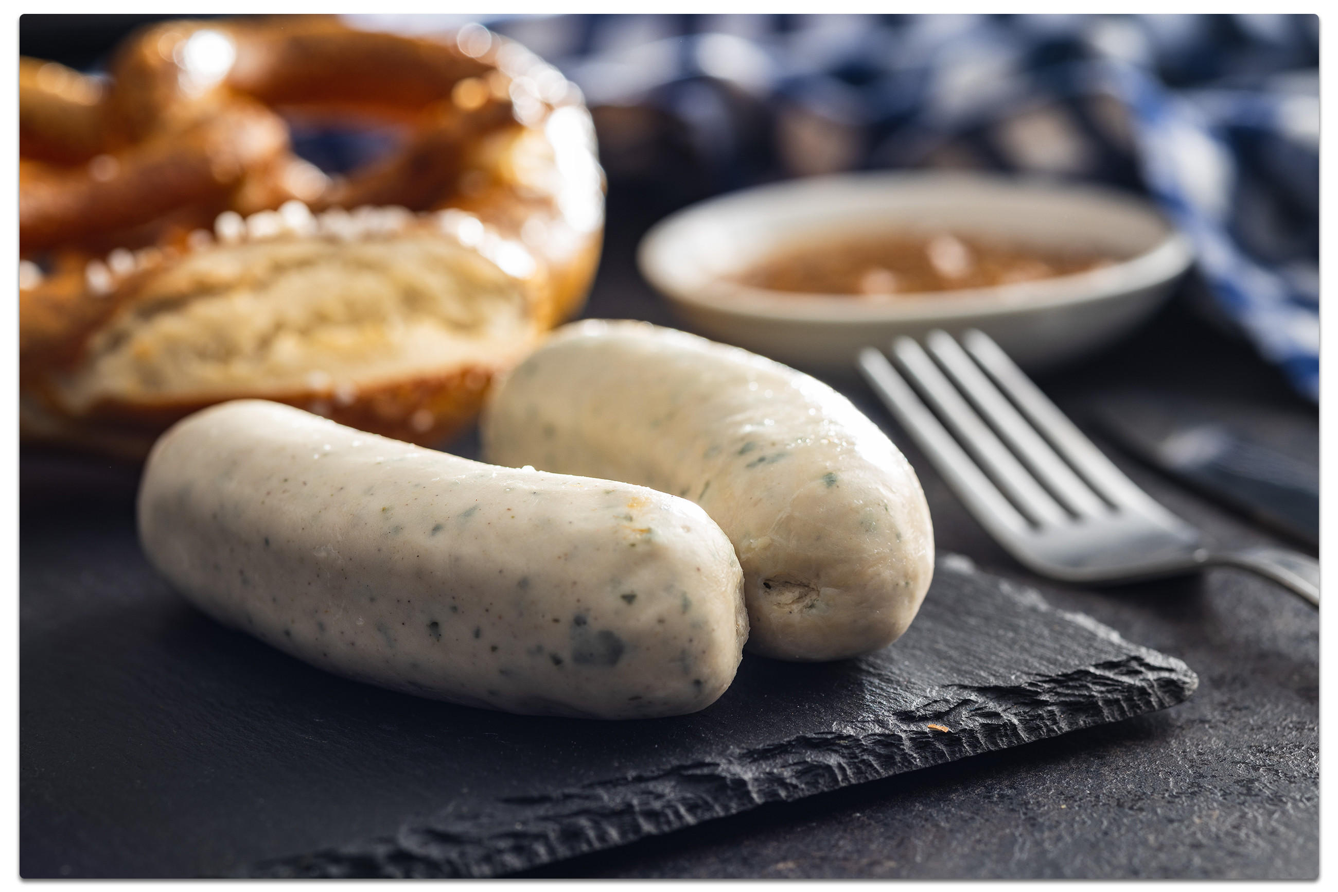 Zwei Weisswürste und eine Brezel im Hintergrund auf einem Schieferbrett, daneben ein Messer und eine Gabel, passend zum Thema Oktoberfest zuhause.