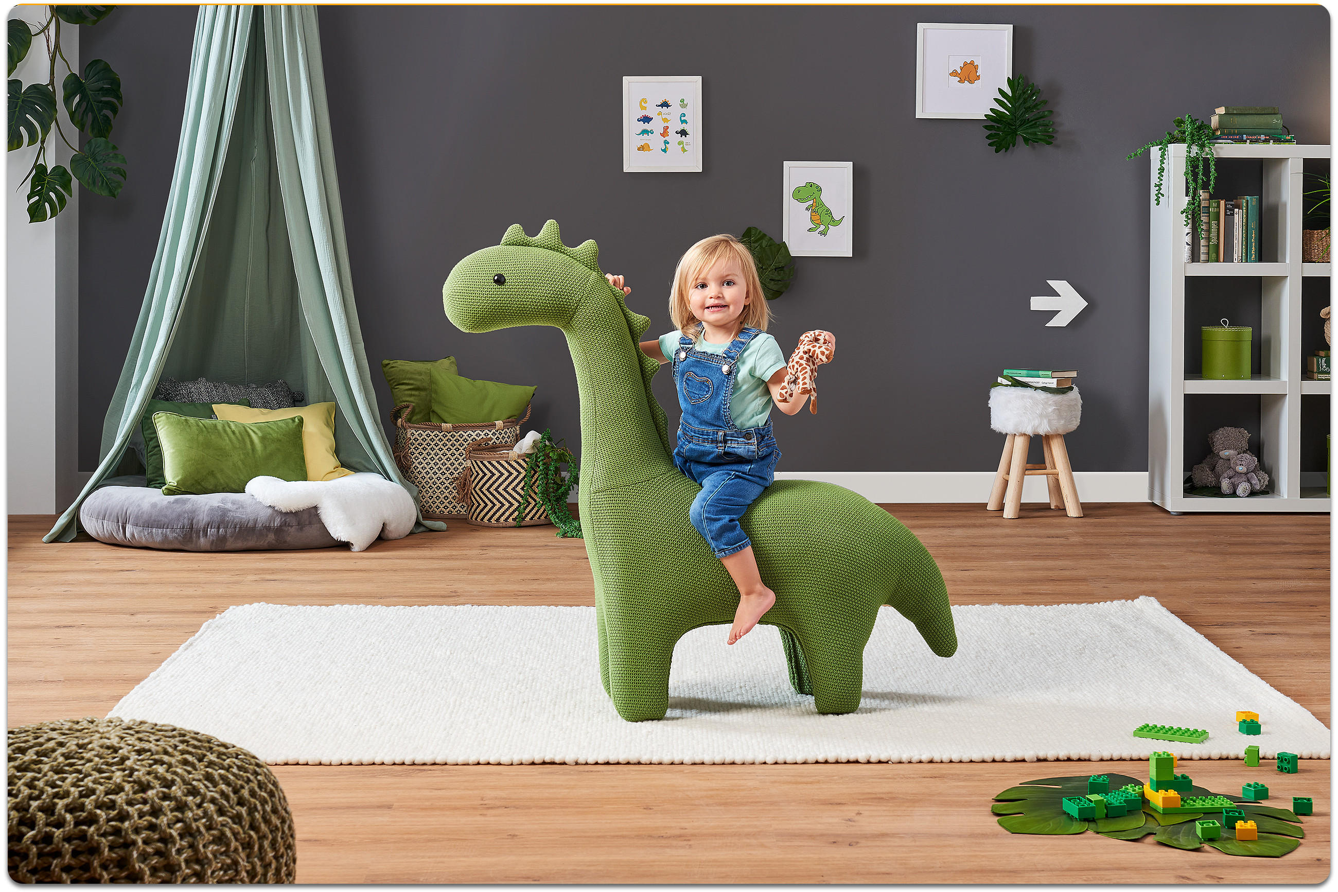 Kind sitzt auf großem grünen Dinosaurier-Kuscheltier in hellem Wohnzimmer mit Teppich und Pflanzen.