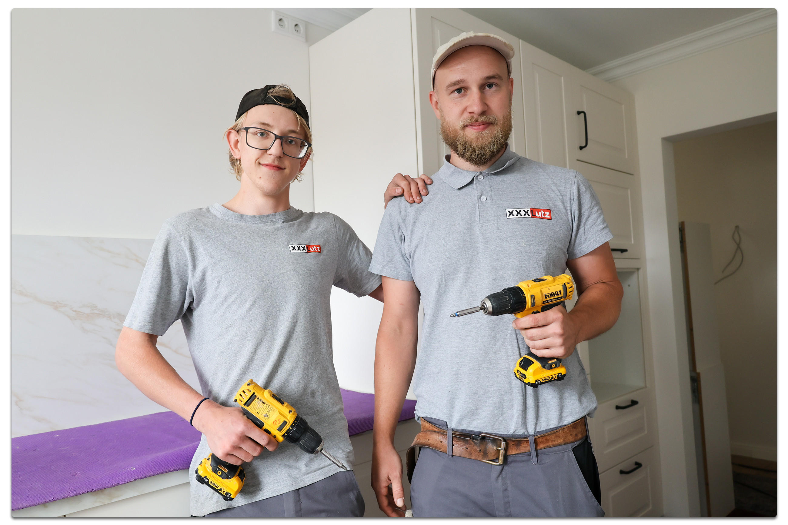 Zwei Männer in grauen T-Shirts mit dem Logo 'XXXL LUTZ' stehen in einer Küche. Beide halten jeweils einen gelben Akkuschrauber in der Hand