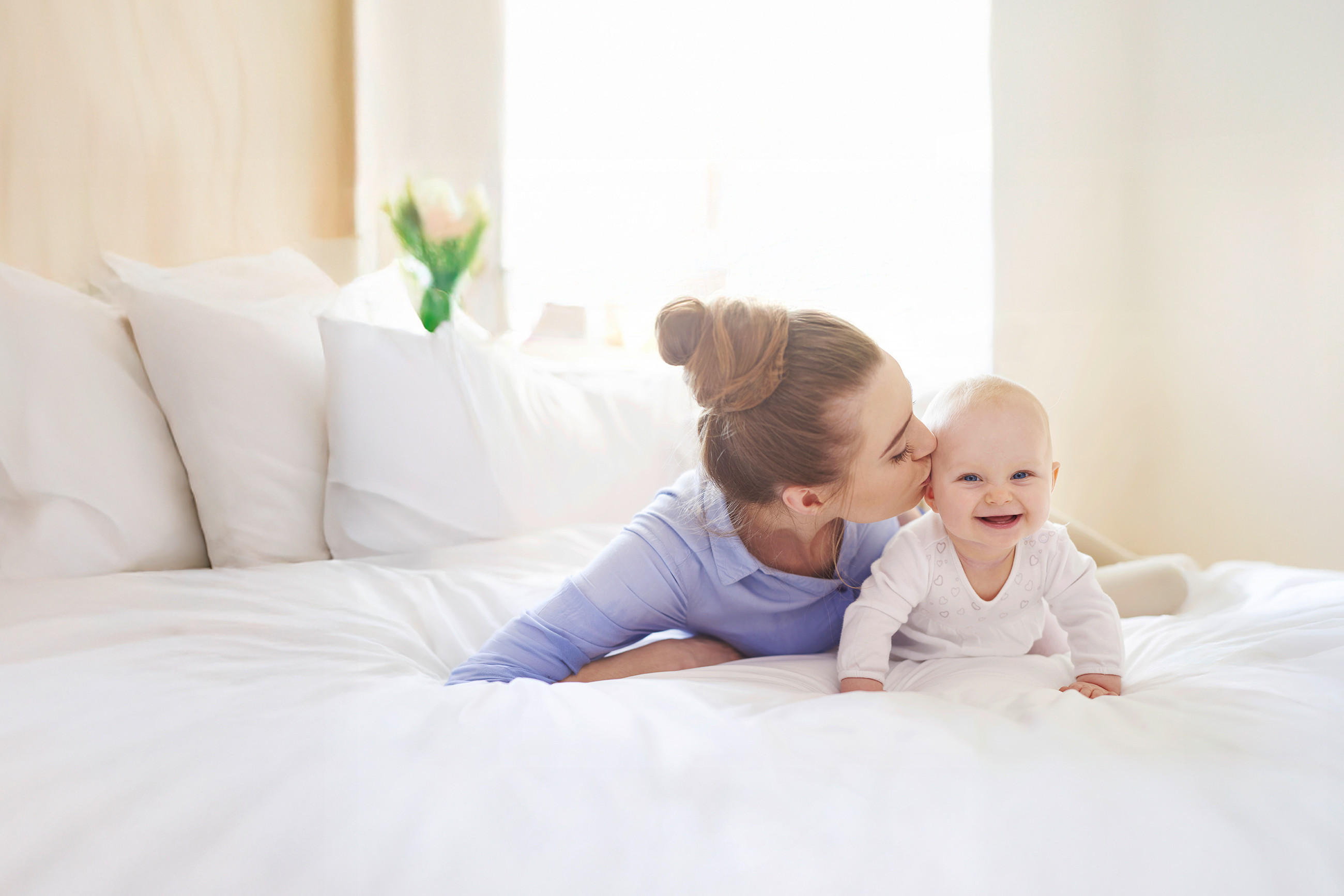 Ein erwachsener Mensch liegt auf einem weißen Bett und lehnt sich zu einem Baby, das aufrecht vor ihm sitzt. Der Raum wirkt hell und freundlich durch das natürliche Licht und die weißen Kissen.