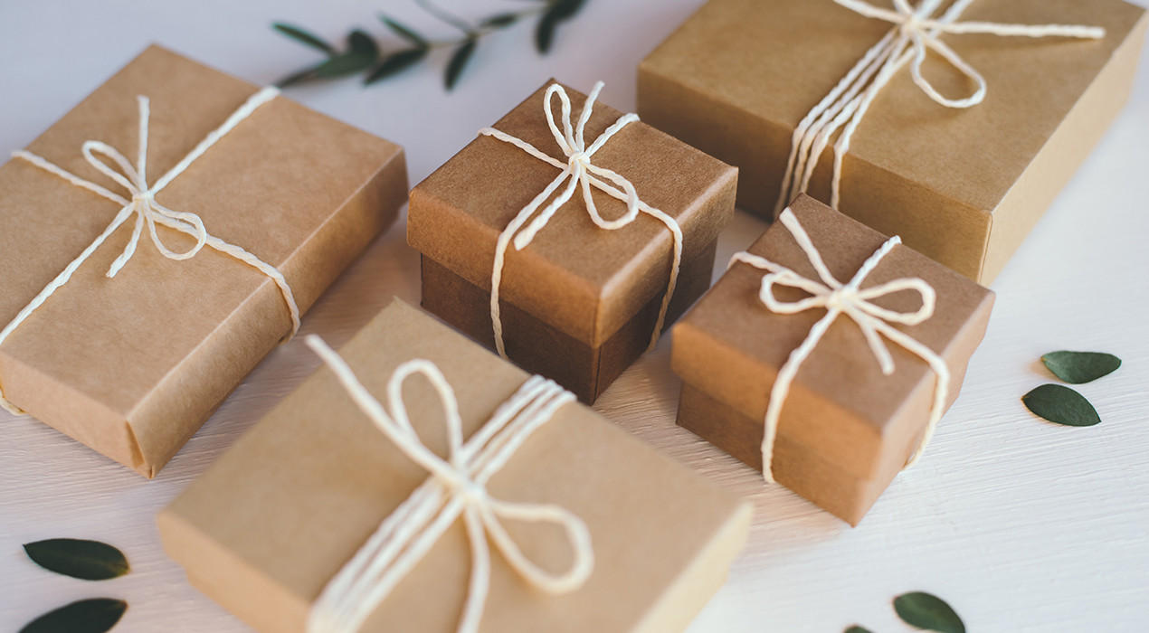 Mehrere braune Geschenkboxen mit weißer Schnur auf hellem Holzuntergrund, daneben grüne Blätter.