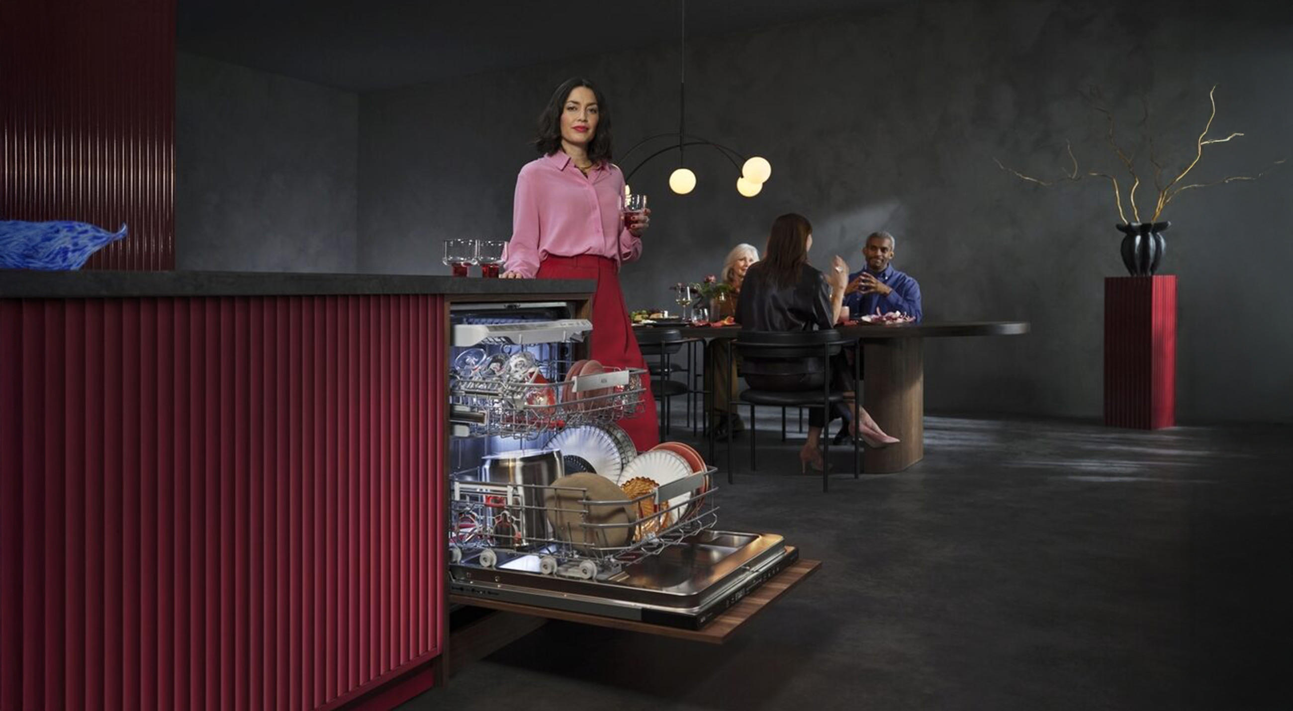 Eine Frau steht vor einem offenen Geschirrspüler von AEG, während im Hintergrund die Gäste dinieren.