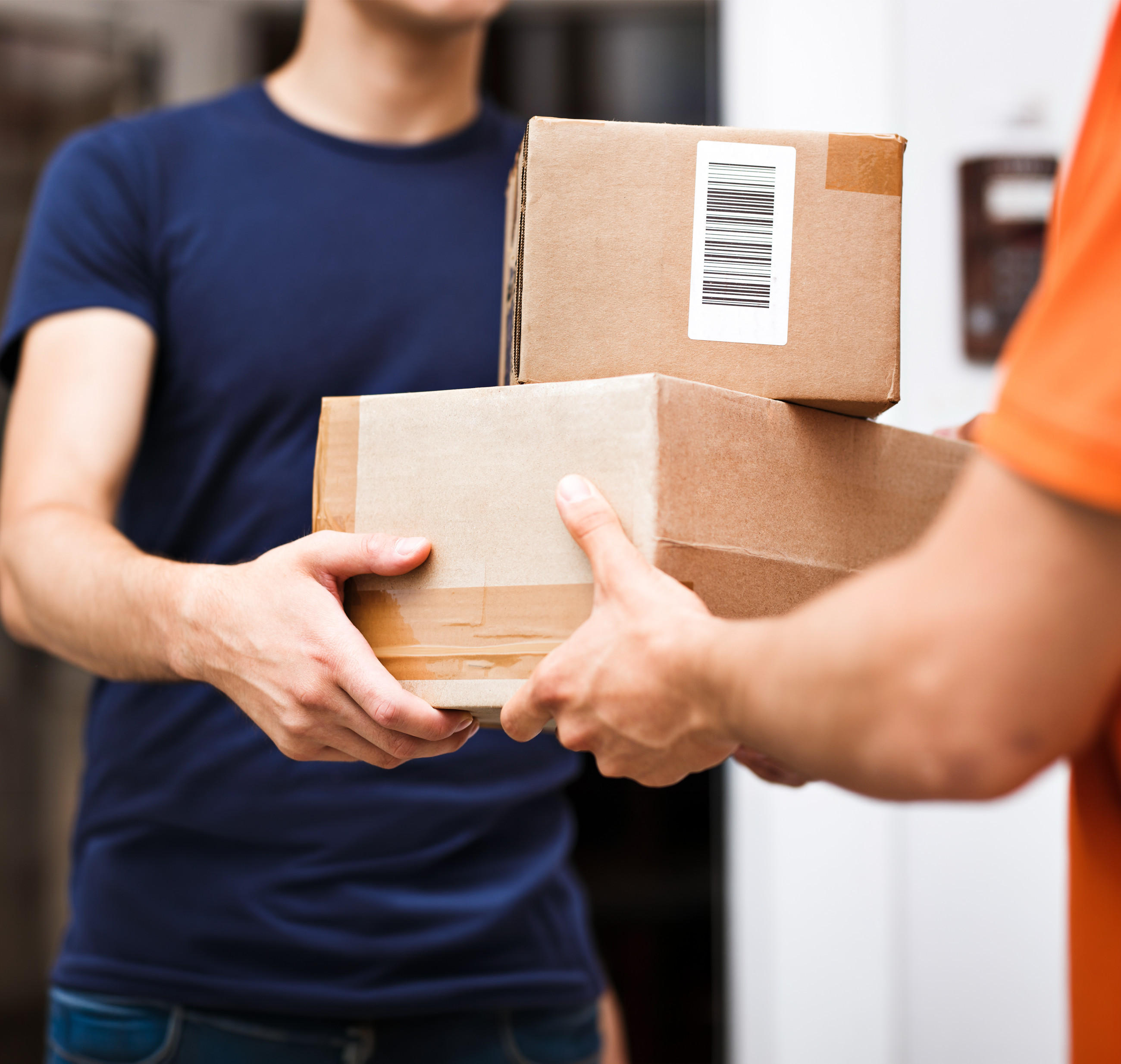  Eine Person trägt ein dunkelblaues T-Shirt, die andere ein orangefarbenes T-Shirt. Die Person im orangefarbenen T-Shirt hält zwei braune Pakete, eines davon mit einem deutlich sichtbaren Barcode. Die Pakete werden der anderen Person übergeben.