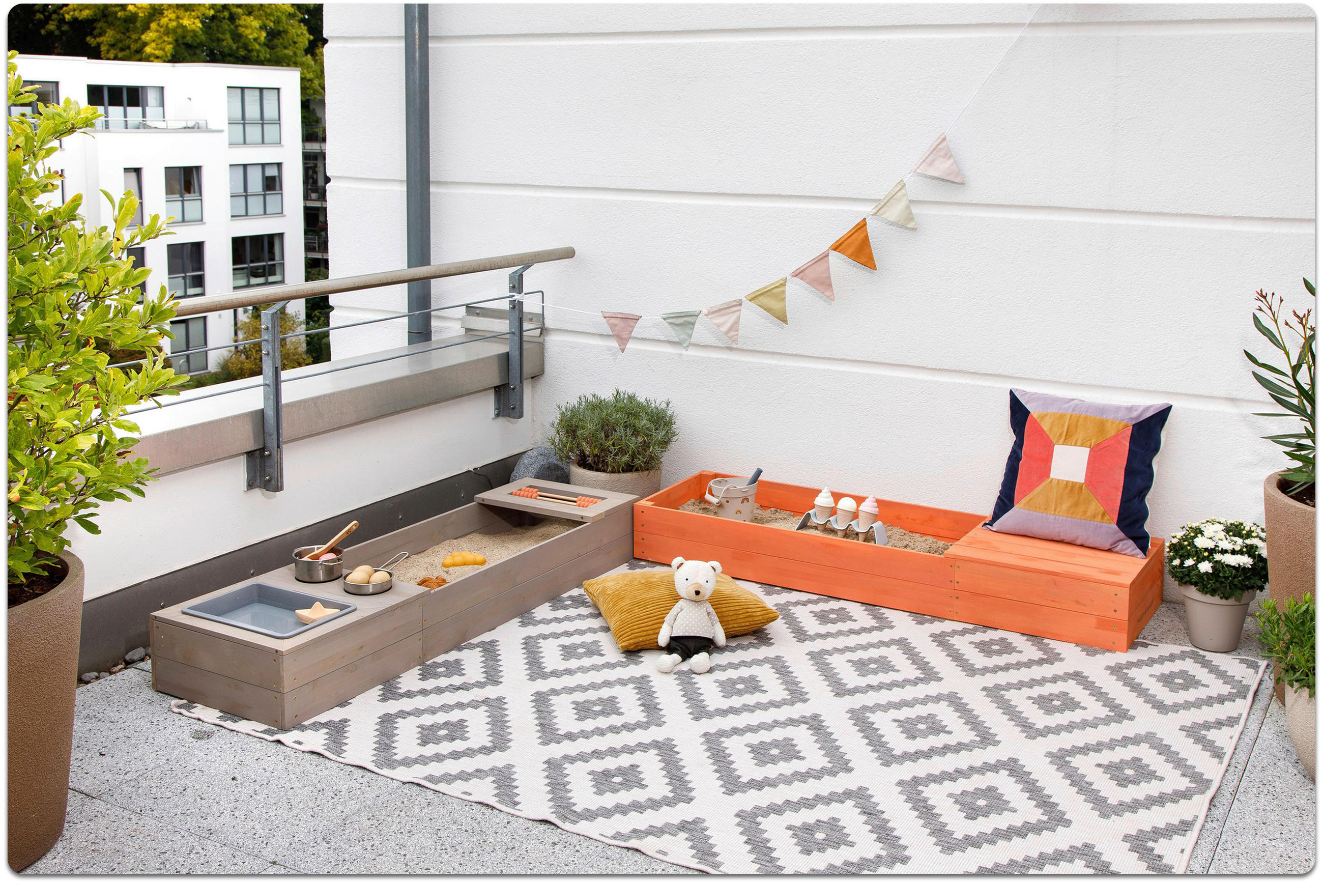 Balkon mit grauem und orangefarbenem Sandkasten, einem gemusterten Teppich, einem Teddybär auf einem gelben Kissen und einer Wimpelkette an der weißen Wand.