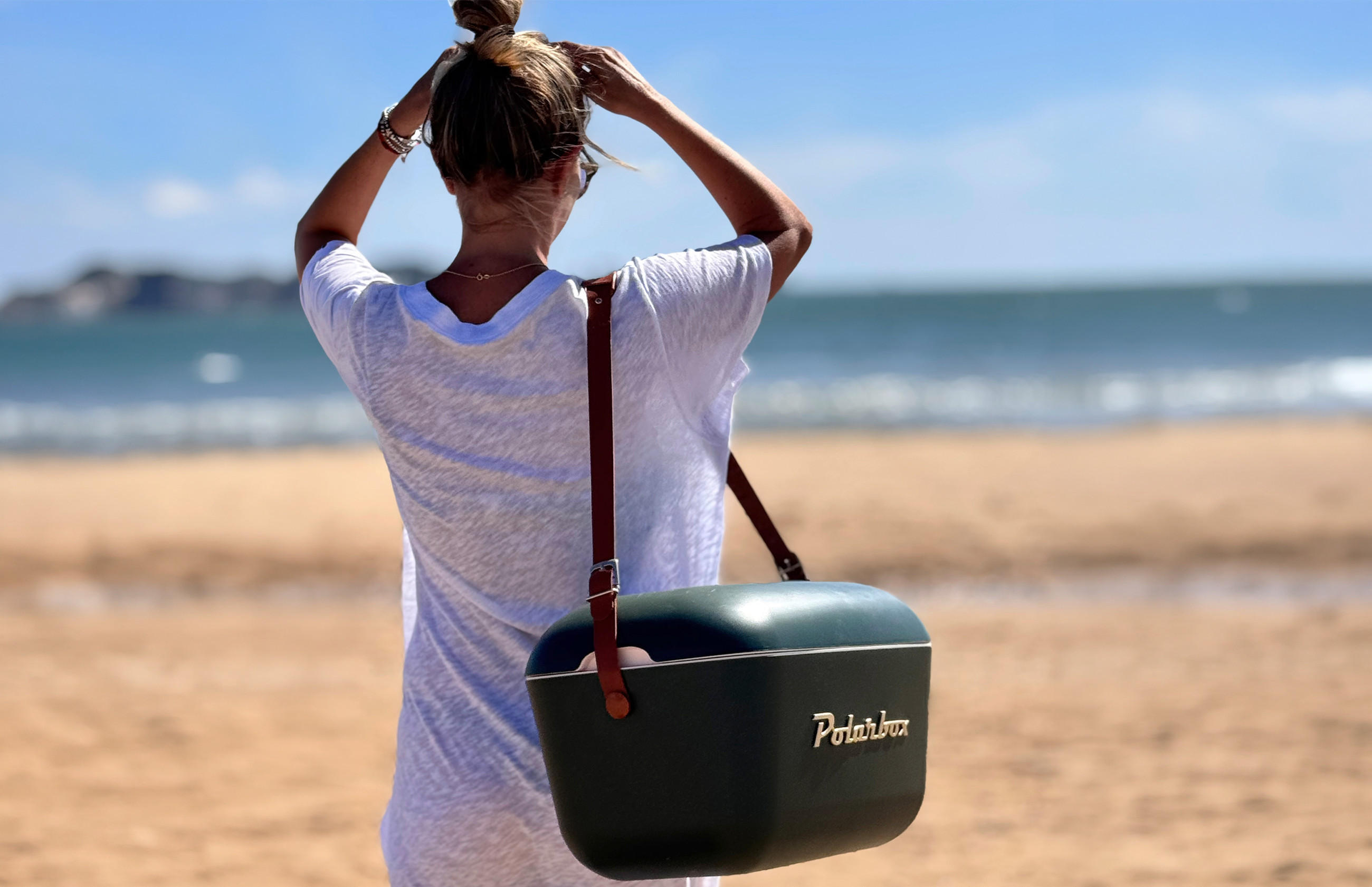 Frau mit hochgestecktem Haar und weißem T-Shirt steht am Strand und blickt aufs Meer, trägt eine grüne Kühltasche mit braunem Tragegurt und goldener Aufschrift