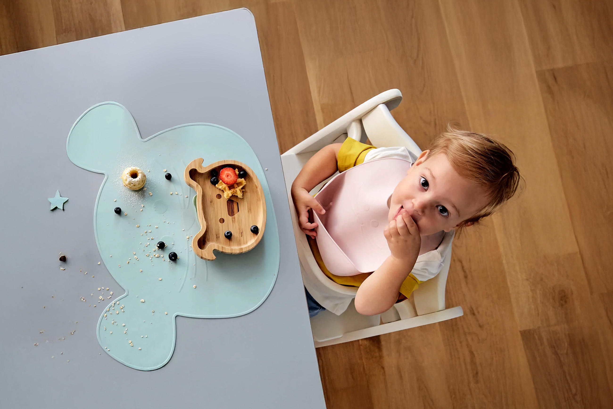 Ein Kleinkind sitzt im Hochstuhl an einem Tisch und isst aus einem geteilten Holz-Teller, der auf einer tierförmigen Silikonunterlage steht. Auf dem Tisch liegen verstreute Haferflocken und Beeren.
