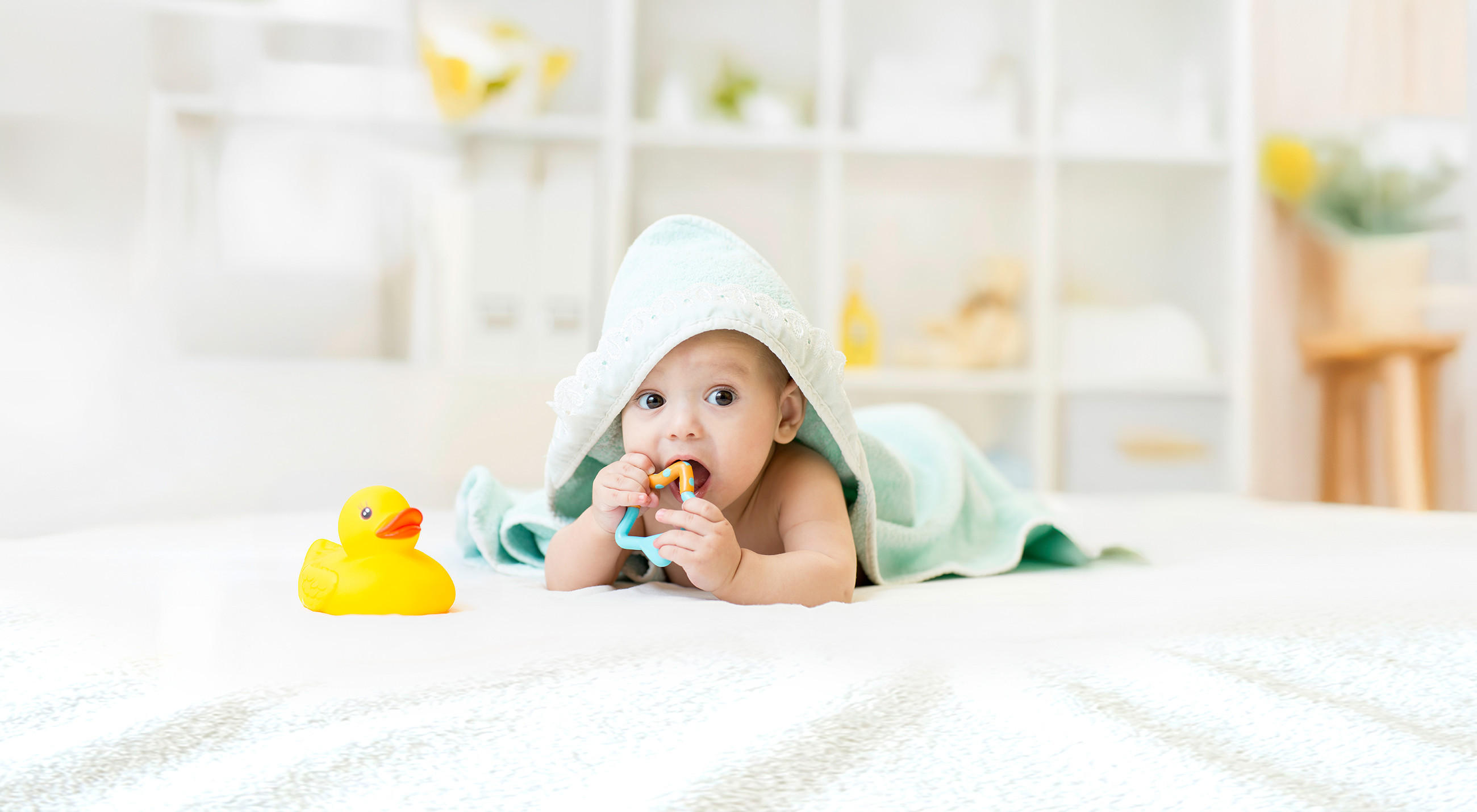 Ein Baby liegt auf dem Bauch auf einem weißen Bett, in ein mintfarbenes Handtuch mit Kapuze gehüllt und hält ein kleines Spielzeug in der Hand. Daneben steht eine gelbe Badeente, und der Hintergrund zeigt ein helles, aufgeräumtes Kinderzimmer.