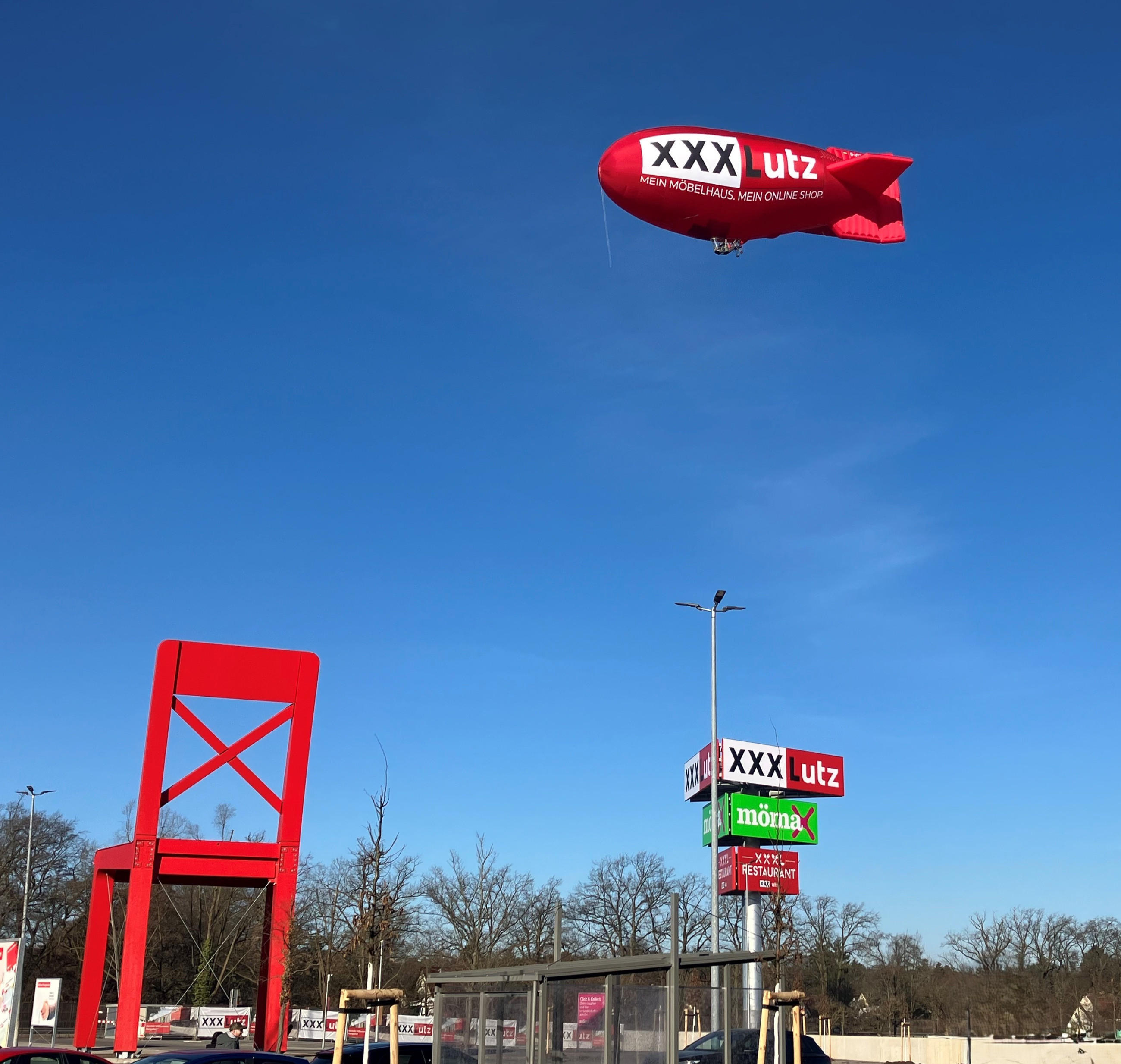 Roter Zeppelin mit XXXLutz-Logo am Himmel. Auf dem Boden befindet sich der große XXXLutz-Stuhl und rechts davon Mastenschilder mit Logo von XXXLutz, Mömax und dem Restaurant.