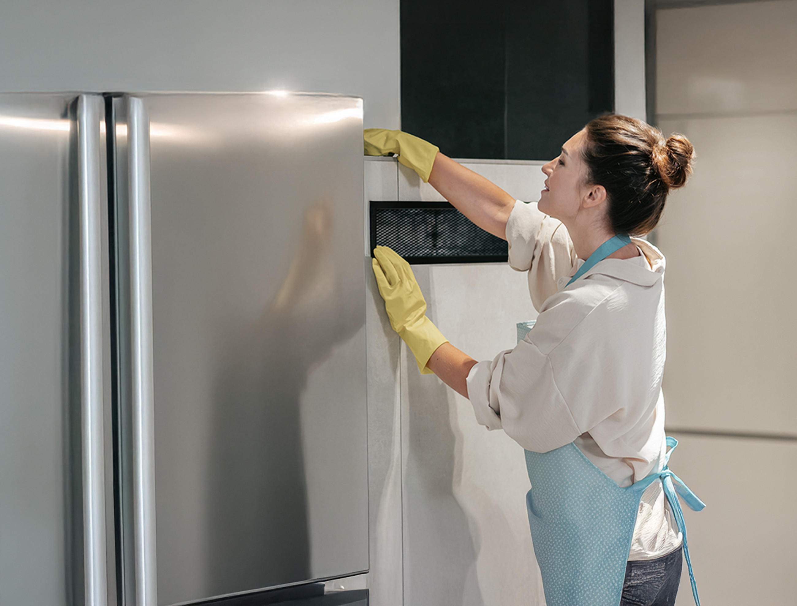 Eine Frau in Handschuhen und Schürze steht in moderner Küche und reinigt einen Kühlschrank von außen.