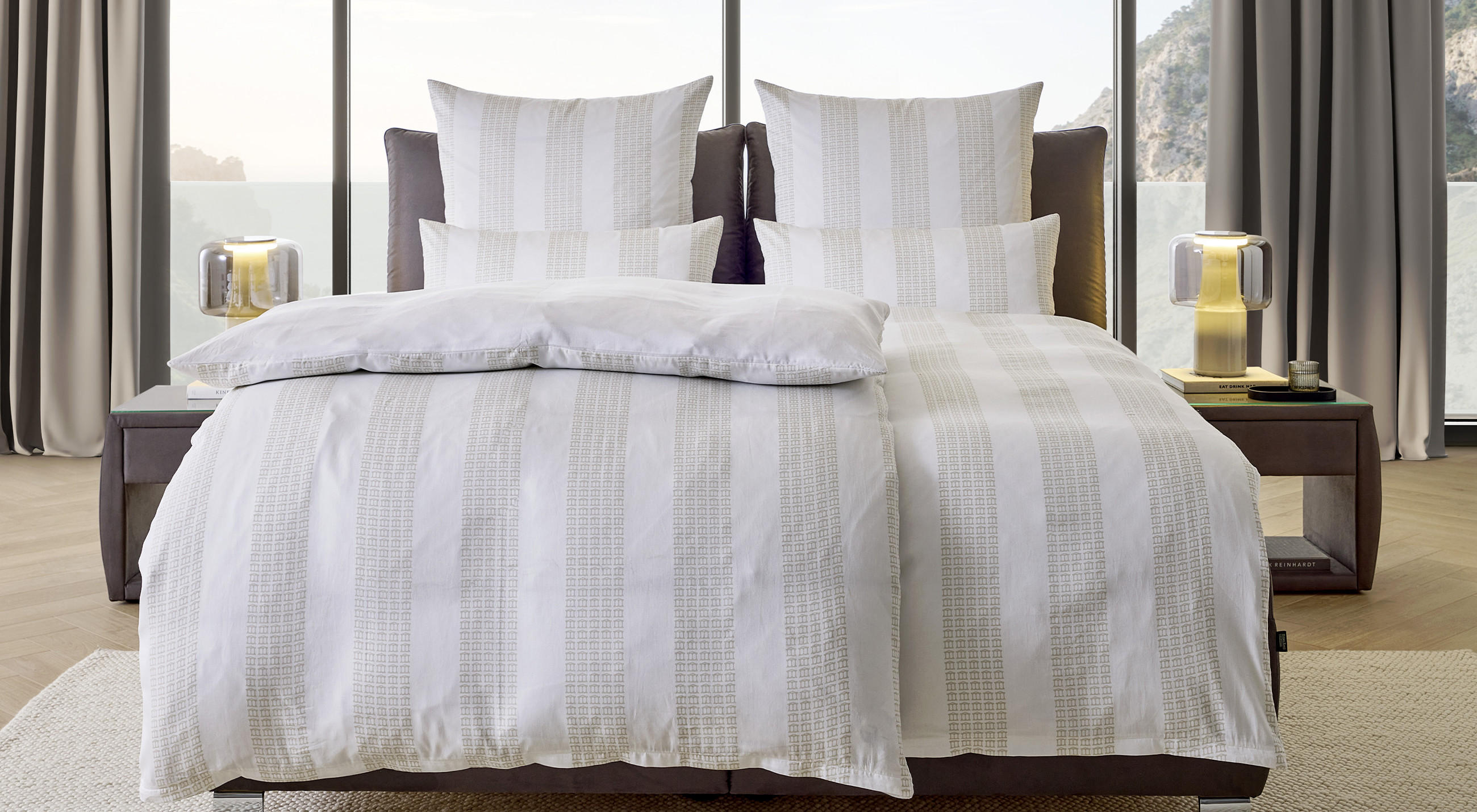 Modernes Schlafzimmer mit großem Bett in Weiß- und Beigetönen, dekoriert mit gestreifter Bettwäsche und mehreren Kissen. Rechts und links Nachttische mit Lampen, im Hintergrund große Fenster mit Berg- und Meerblick