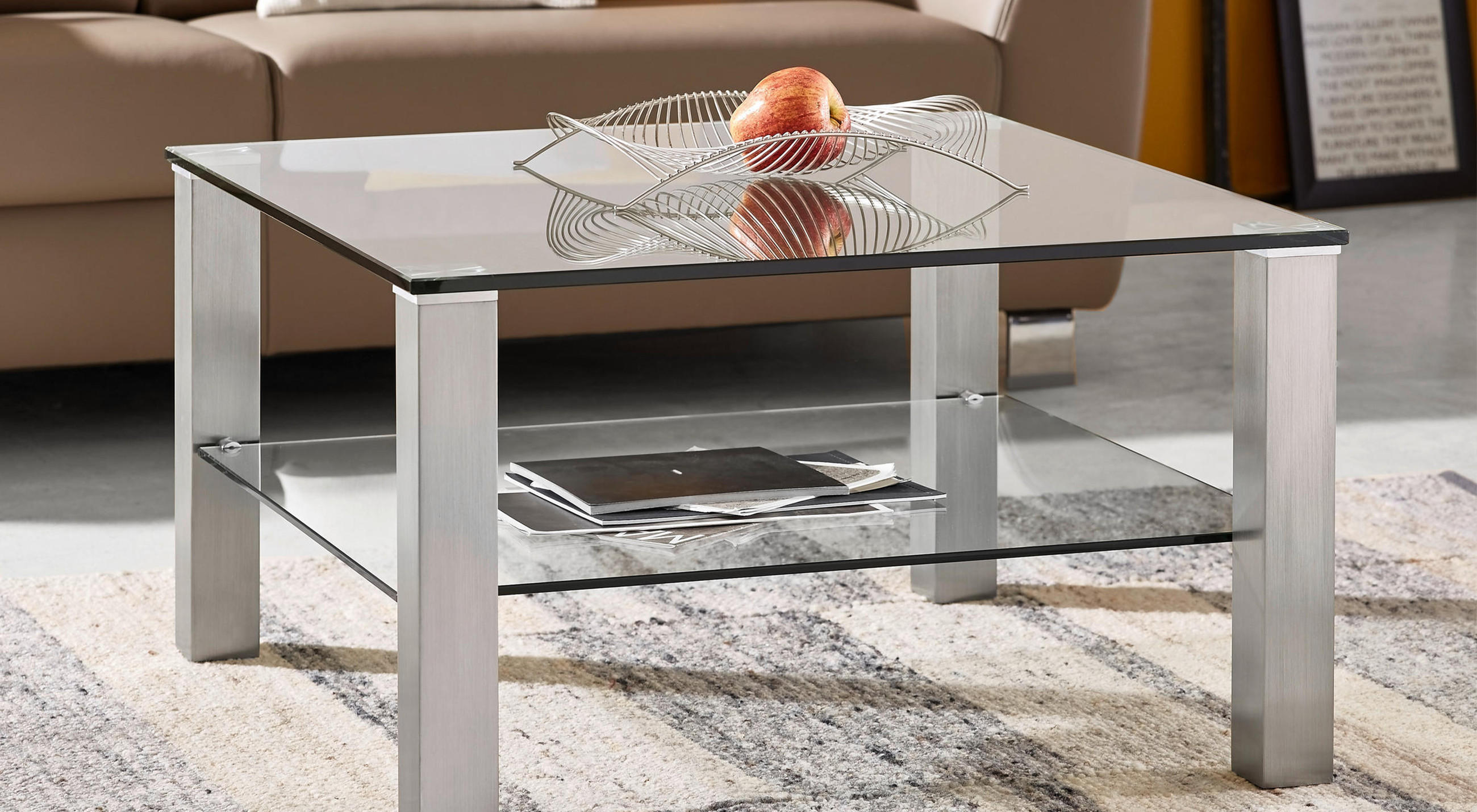 Quadratischer Couchtisch mit Glasplatte und Unterboden aus Glas, silbernen Metallbeinen und dekorativer Drahtschale mit Apfel steht auf gemustertem Teppich vor braunem Sofa