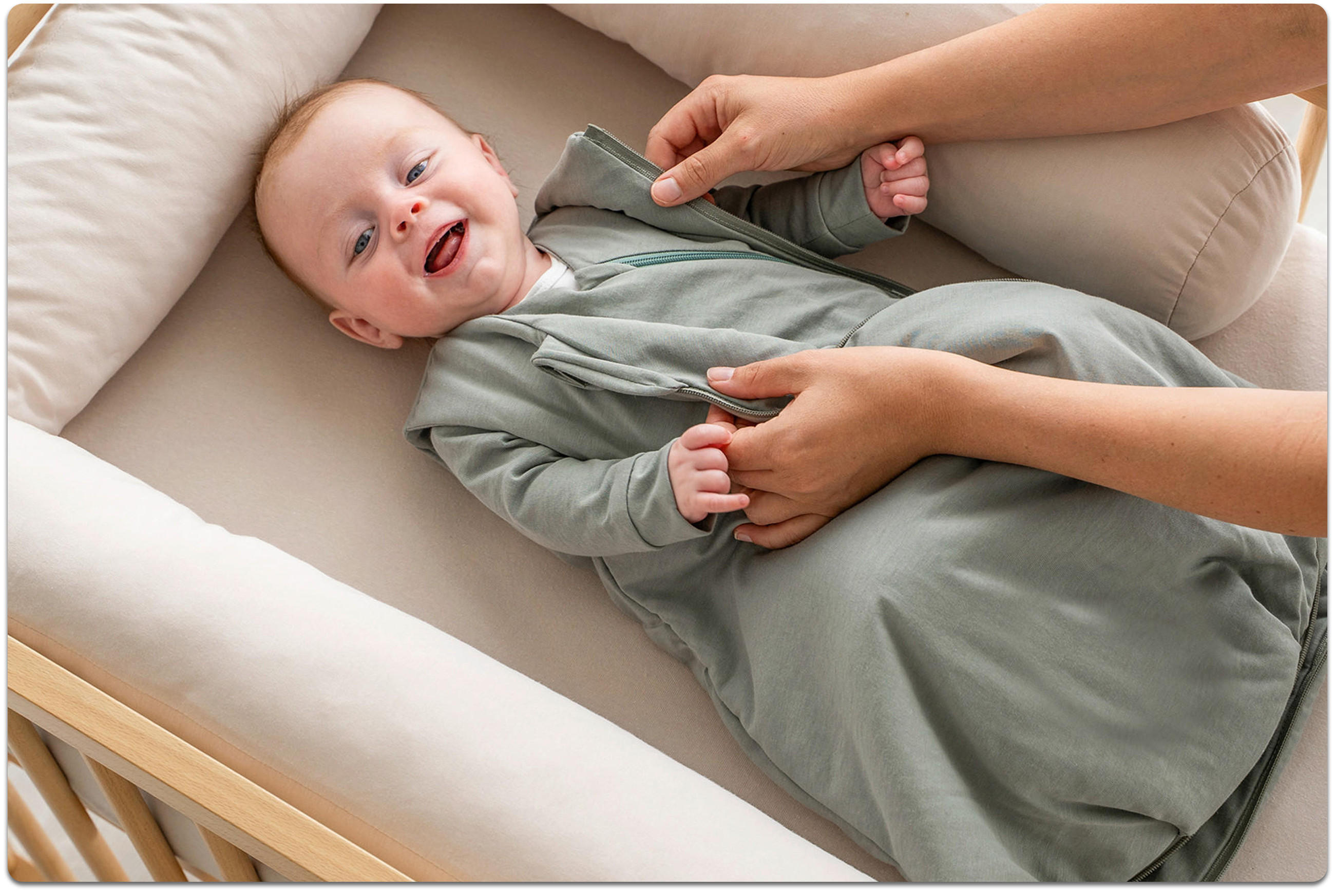 Ein Baby liegt in einem beigen Babynestchen; es trägt einen grünen Body und lächelt. Die Hände eines Elternteils greifen das Baby seitlich und berühren es sanft, um es hochzuheben.