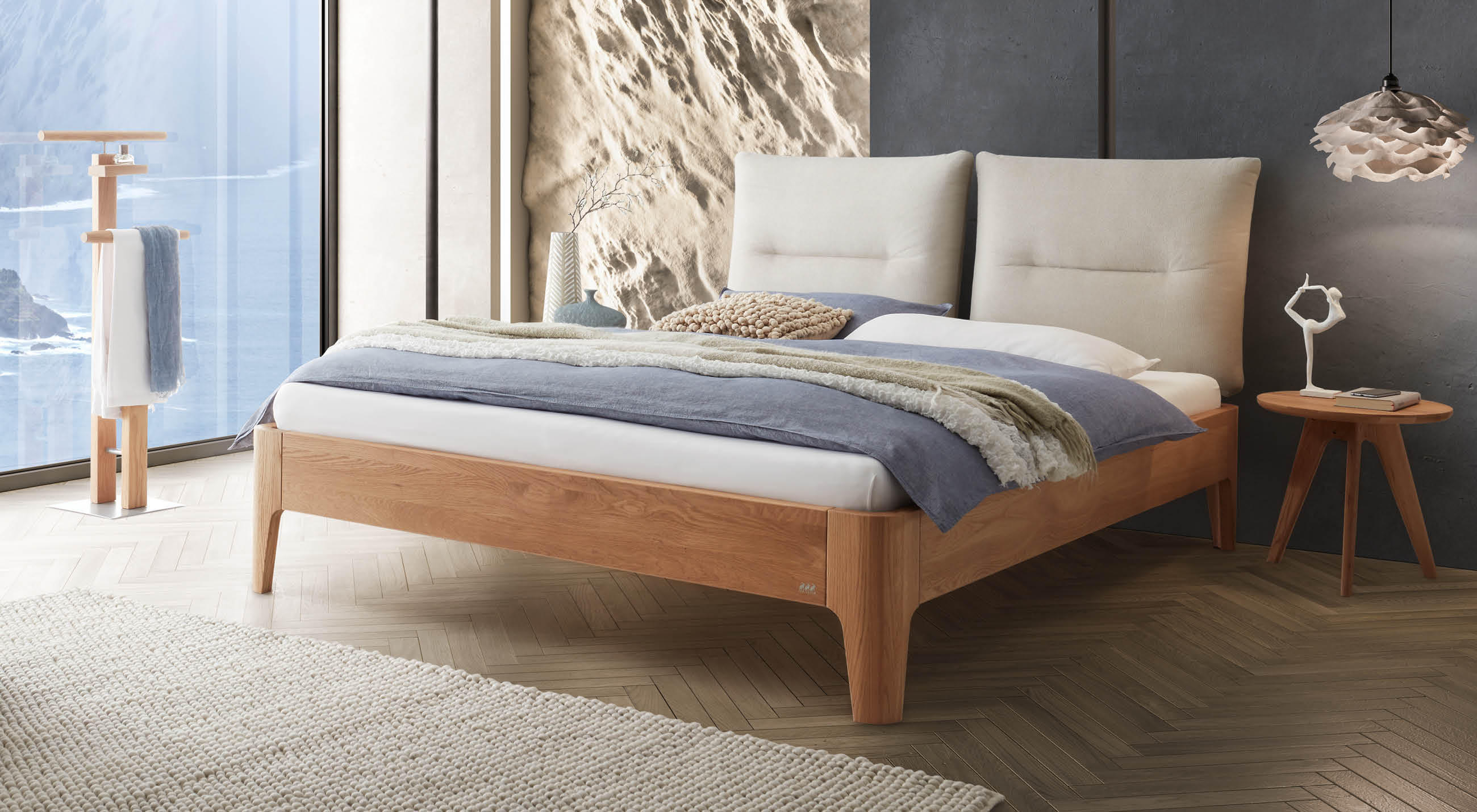Modernes Hasena Bett aus Massivholz mit gepolstertem Doppelkopfteil, natürlicher Bettwäsche und stilvollen Accessoires in einem hellen Schlafzimmer mit großem Fenster.