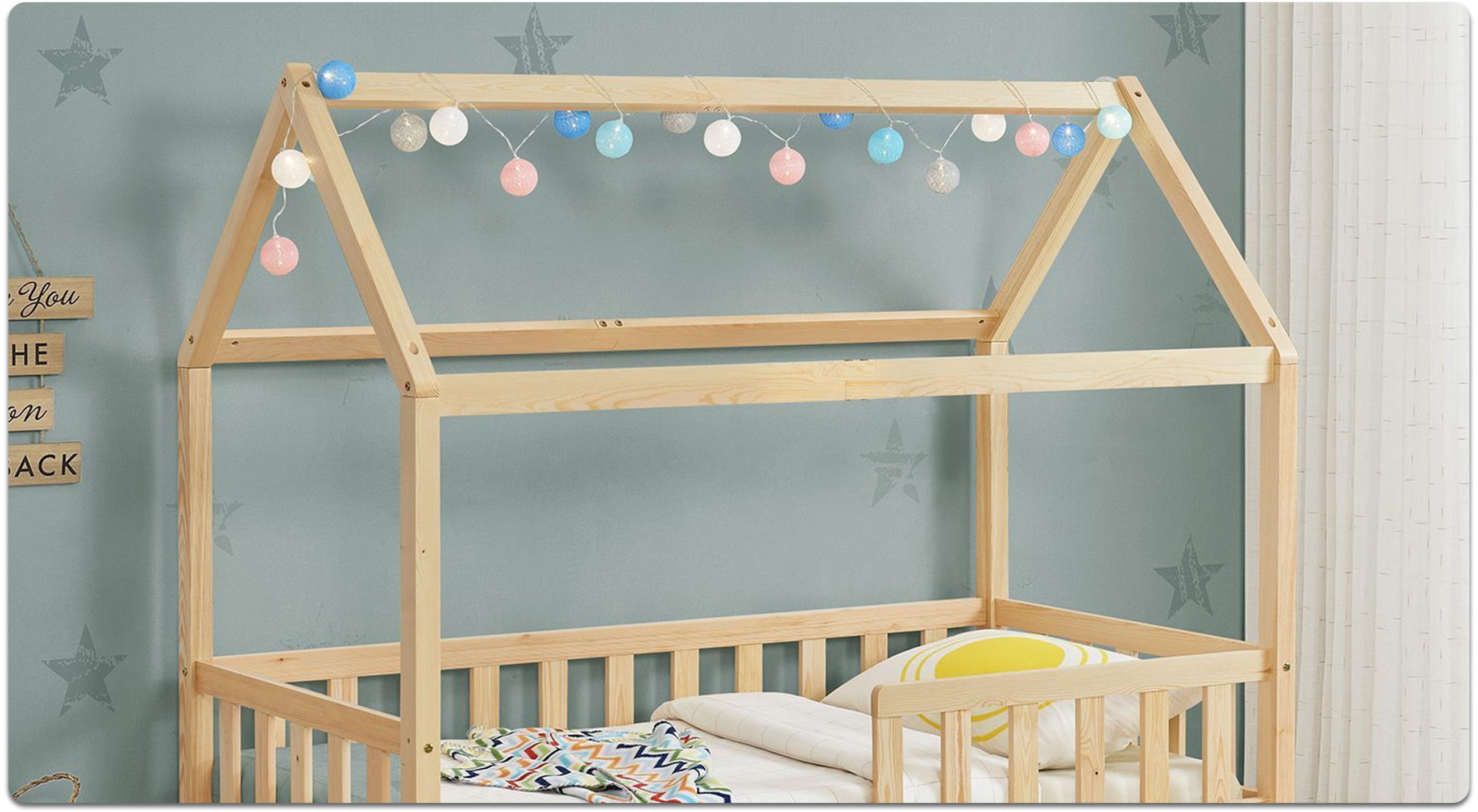 Kinderbett mit Holzrahmen in Hausform und einer Lichterkette aus bunten Kugeln an der oberen Querstrebe vor einer Wand mit Sternenmotiven.