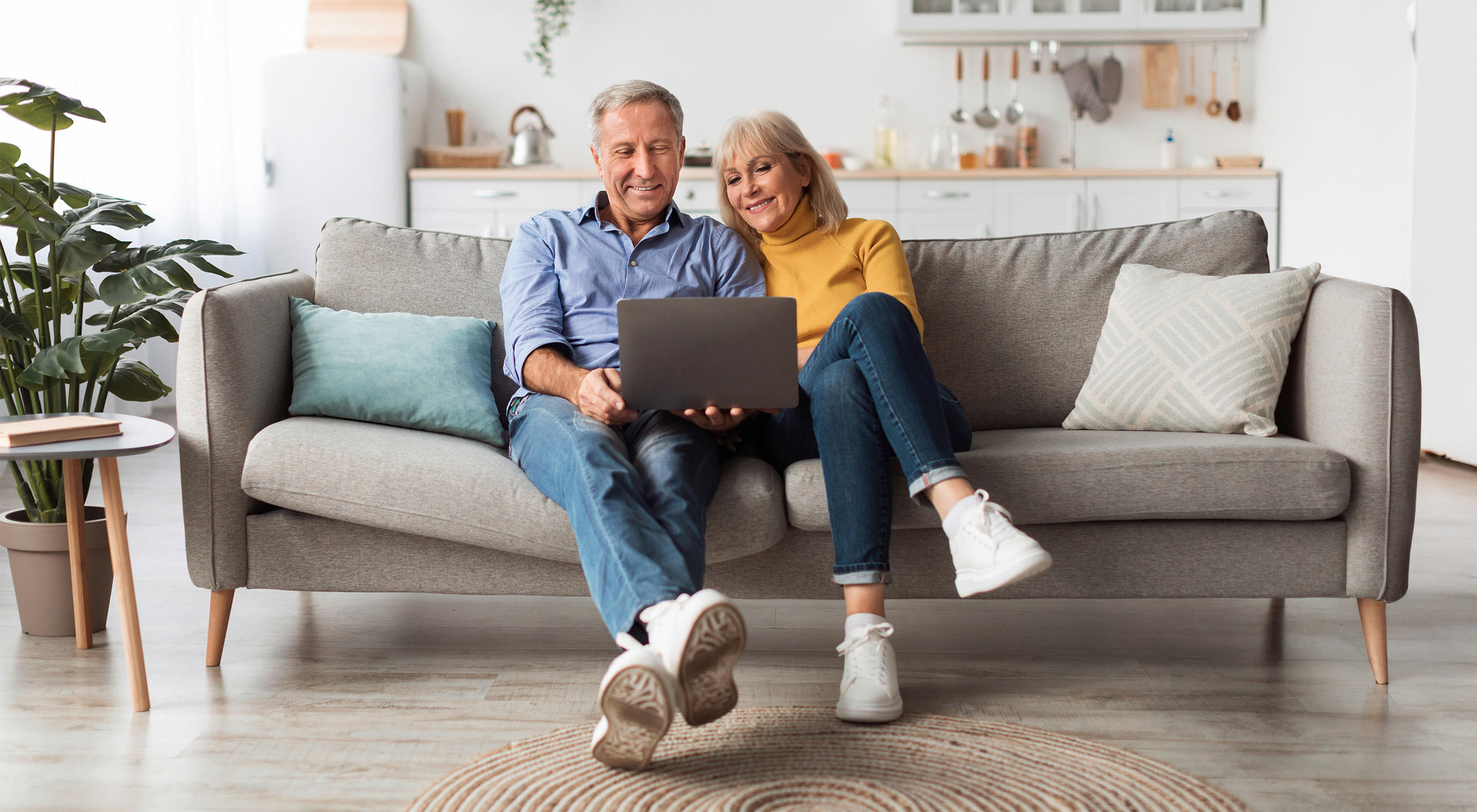 Mann und Frau sitzen bequem auf einem grauen Sofa und planen ihre neue Küche am Laptop. Im Hintergrund ist ihre aktuelle, weiße Küchenzeile zu erkennen..
