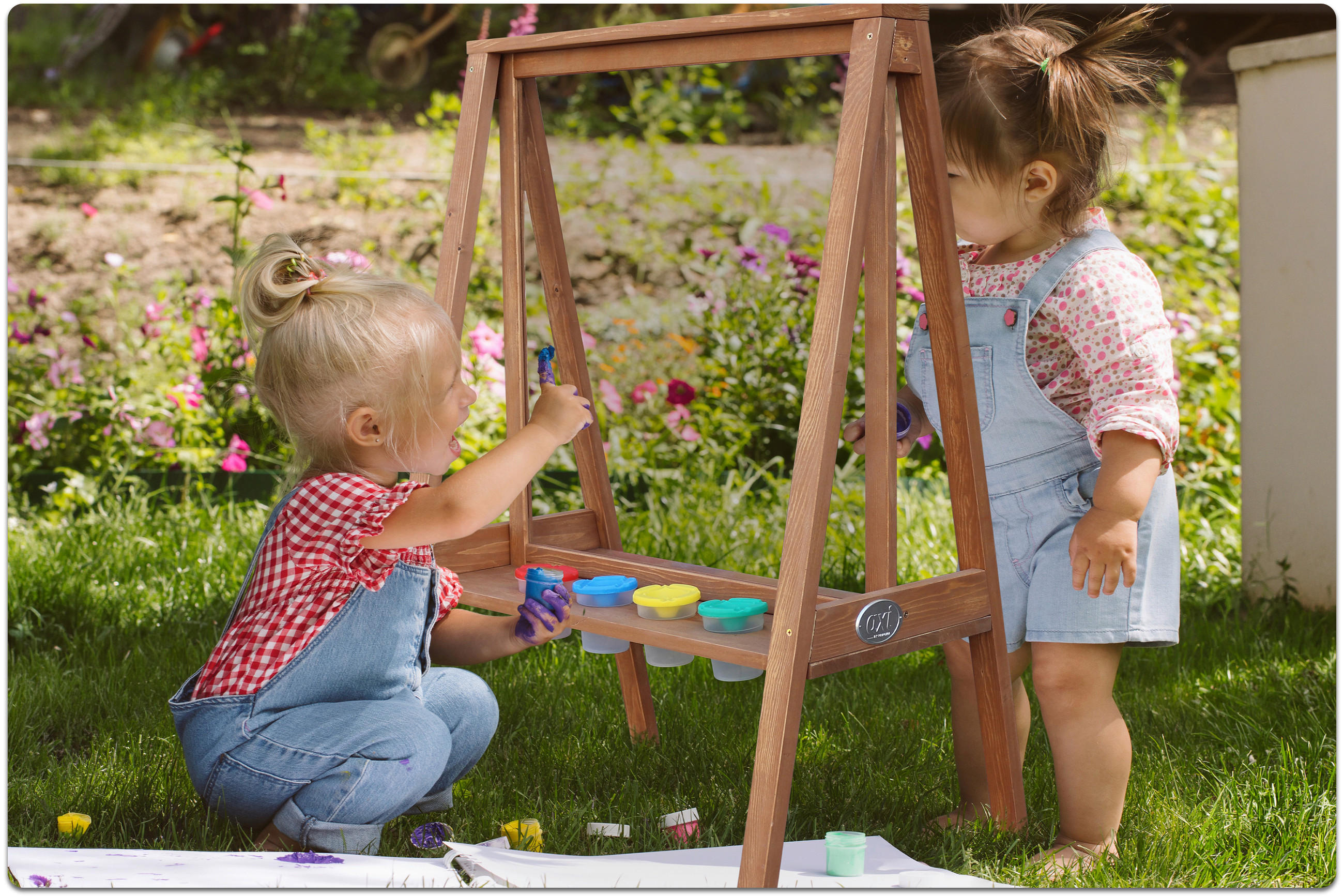 Zwei Kleinkinder mit Latzhosen malen ein Bild mit Farben, die in einer Holzstaffelei im Garten steht.