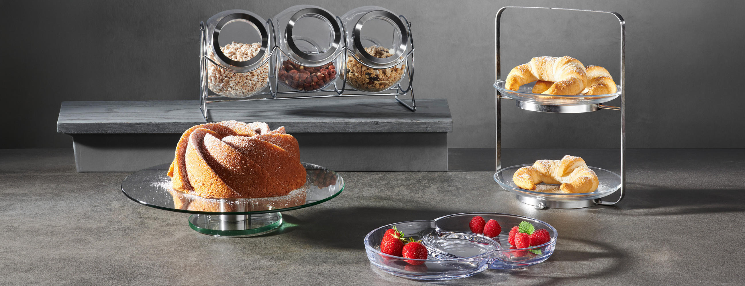Kuchen auf Glasplatte, daneben Glasbehälter mit Nüssen und Trockenfrüchten sowie ein zweistöckiges Gestell mit Croissants und eine Glasplatte mit Erdbeeren und Himbeeren.