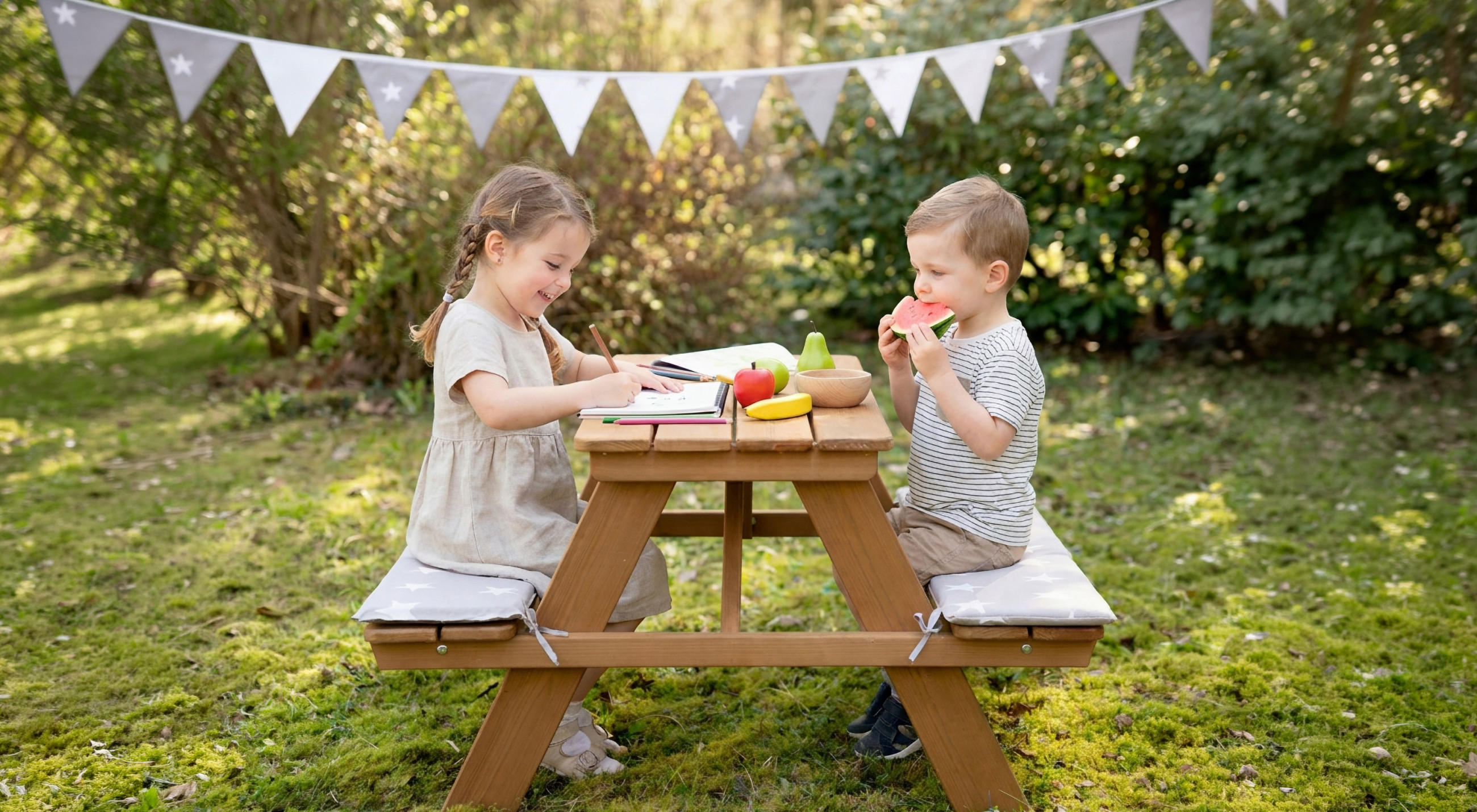 Zwei Kinder sitzen an einem kleinen Holztisch im Garten und essen beziehungsweise malen.
