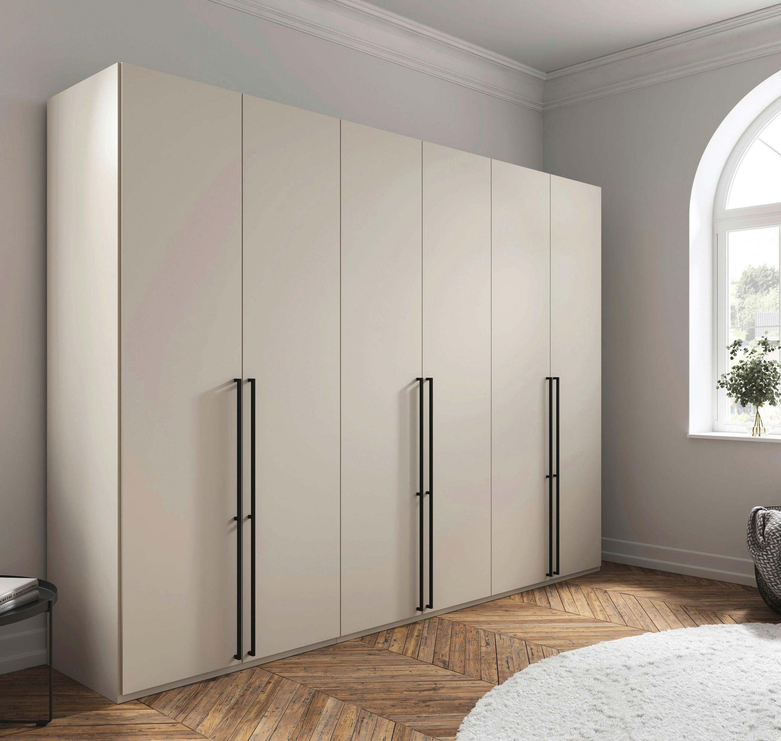 Heller Kleiderschrank mit mehreren Türen und schwarzen Griffen in lichtdurchflutetem Zimmer mit großen Rundbogenfenstern und Holzparkett.