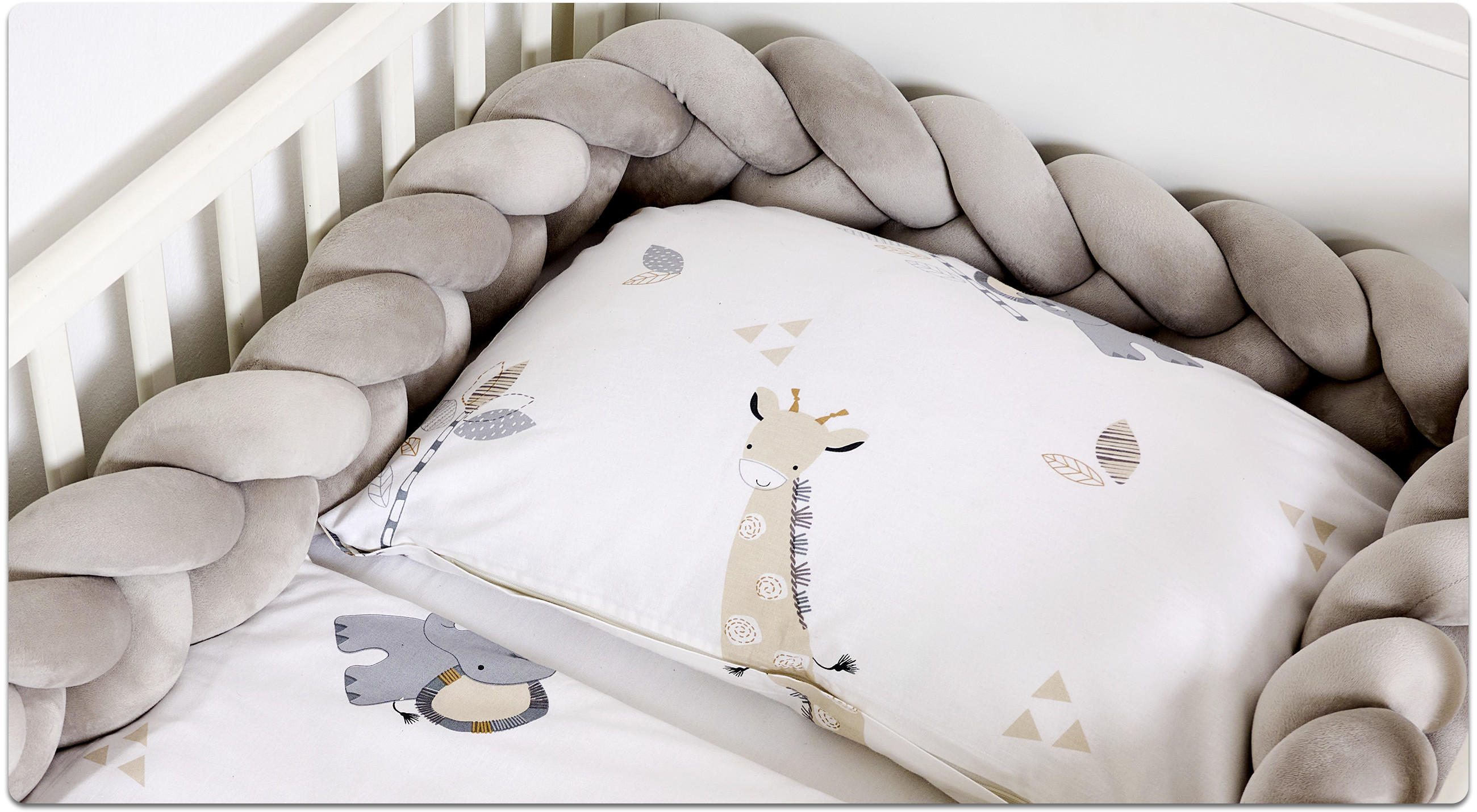 Nahaufnahme eines Babybetts mit weißer Bettwäsche mit Giraffen- und Blättermotiven, umrahmt von einem geflochtenen, gepolsterten Bettschlangen-Rand in Beige.