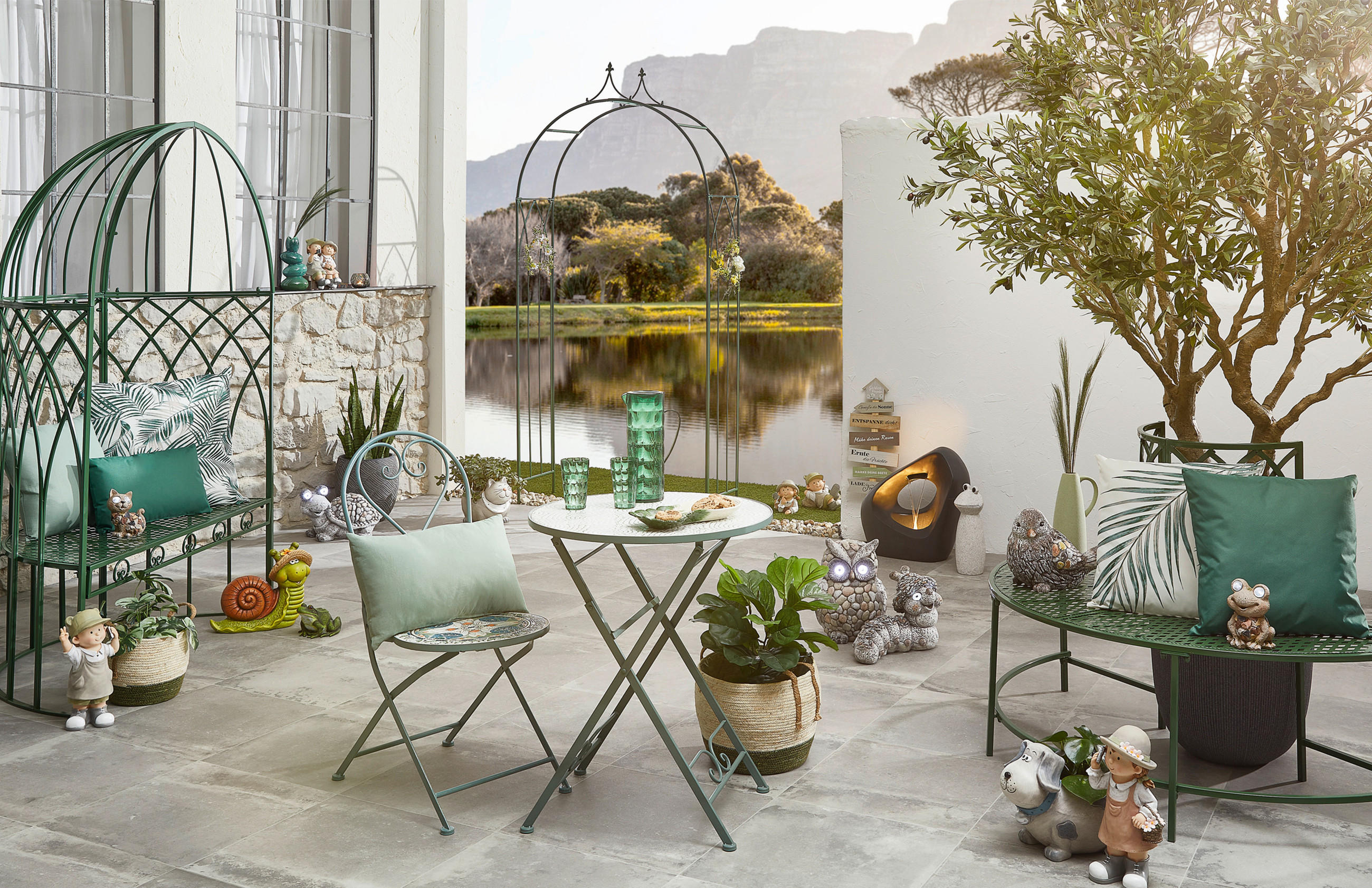 Terrasse mit grün lackierten Metallmöbeln, Kissen, Pflanzen und verschiedenen Gartendekorationen vor einem See mit Bäumen im Hintergrund.