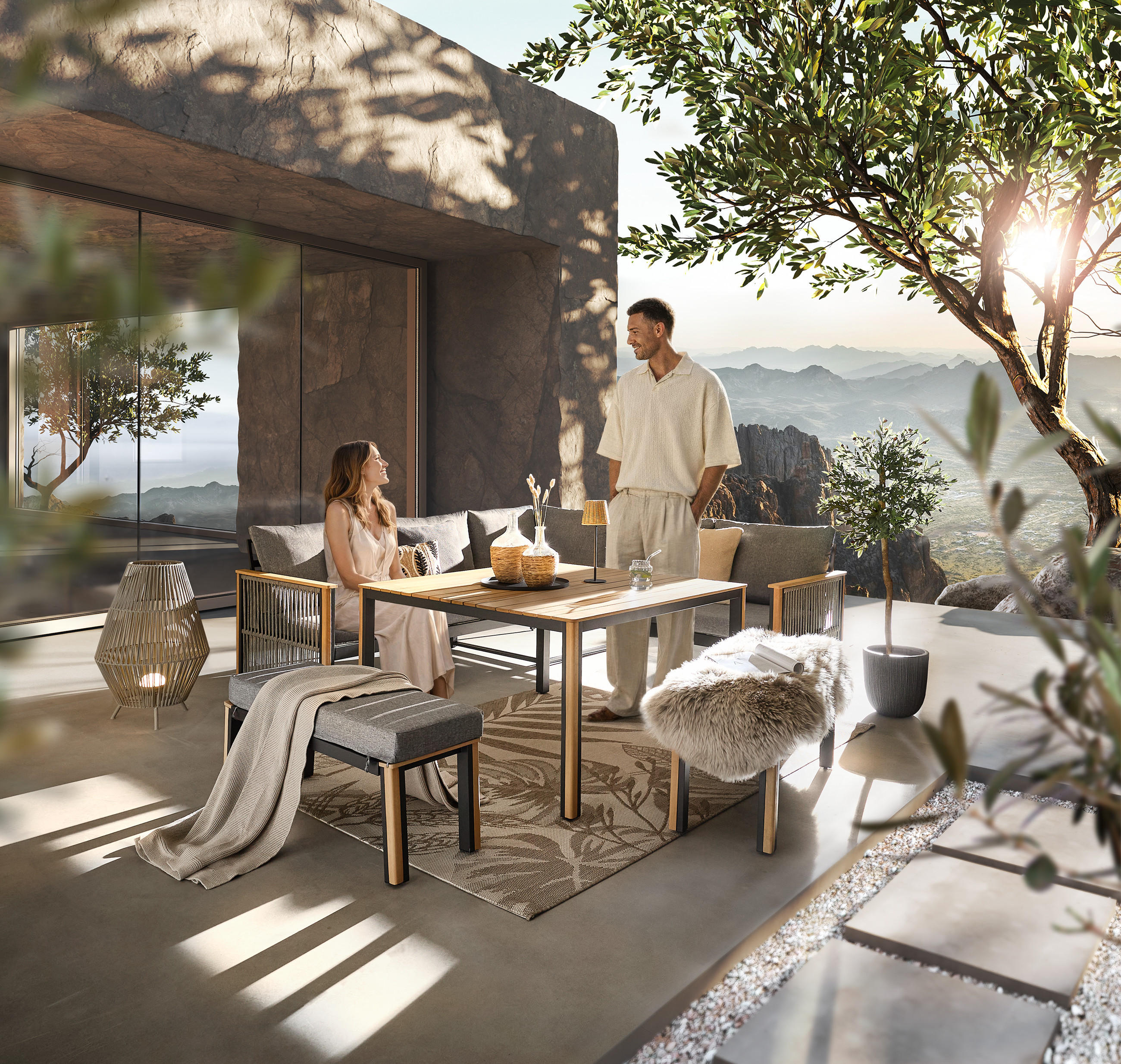 Zwei Personen sitzen und stehen an einem modernen Gartentisch auf einer Terrasse mit Blick auf Berge und Pflanzen.