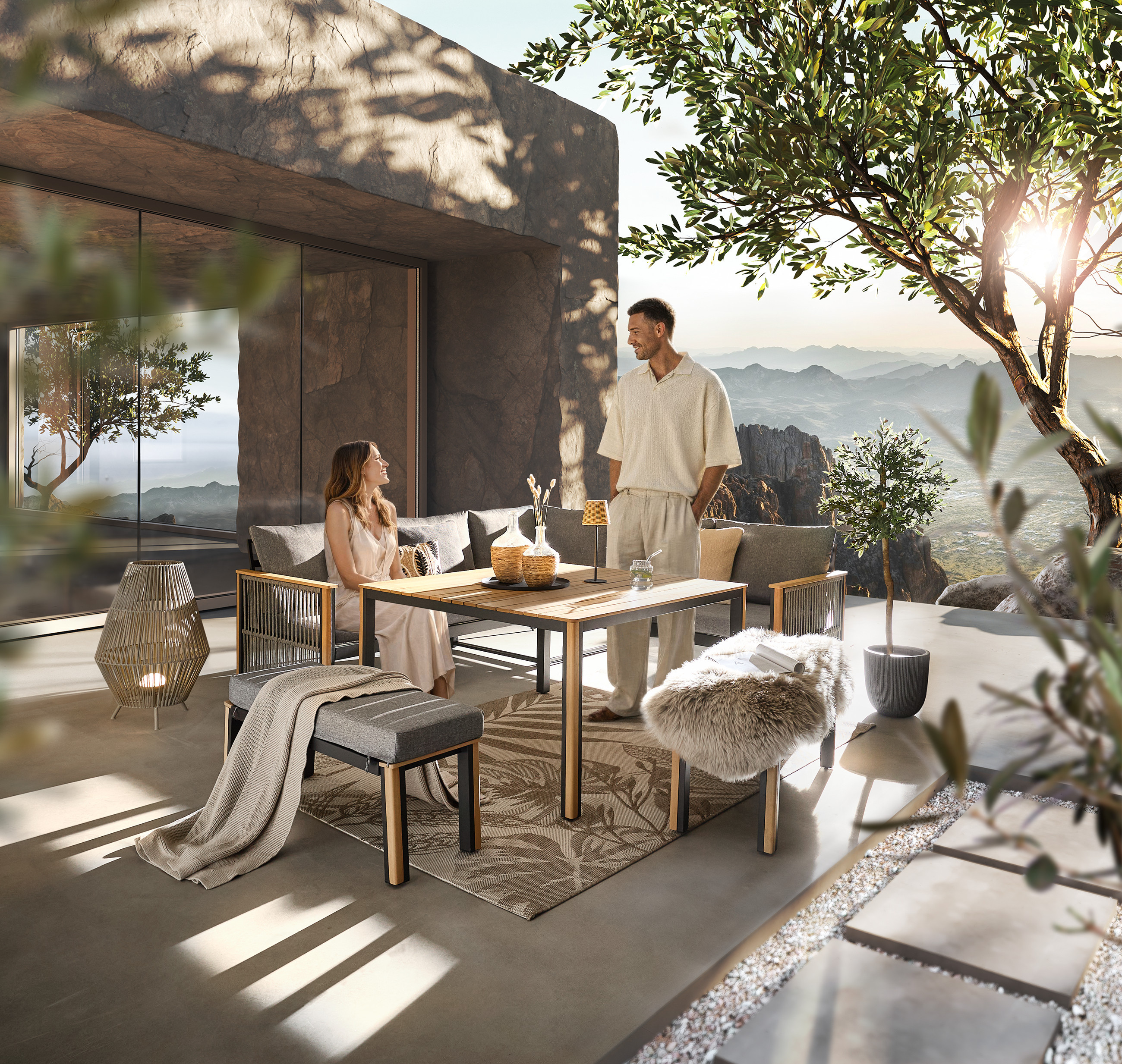 Zwei Personen sitzen und stehen an einem modernen Gartentisch auf einer Terrasse mit Blick auf Berge und Pflanzen.