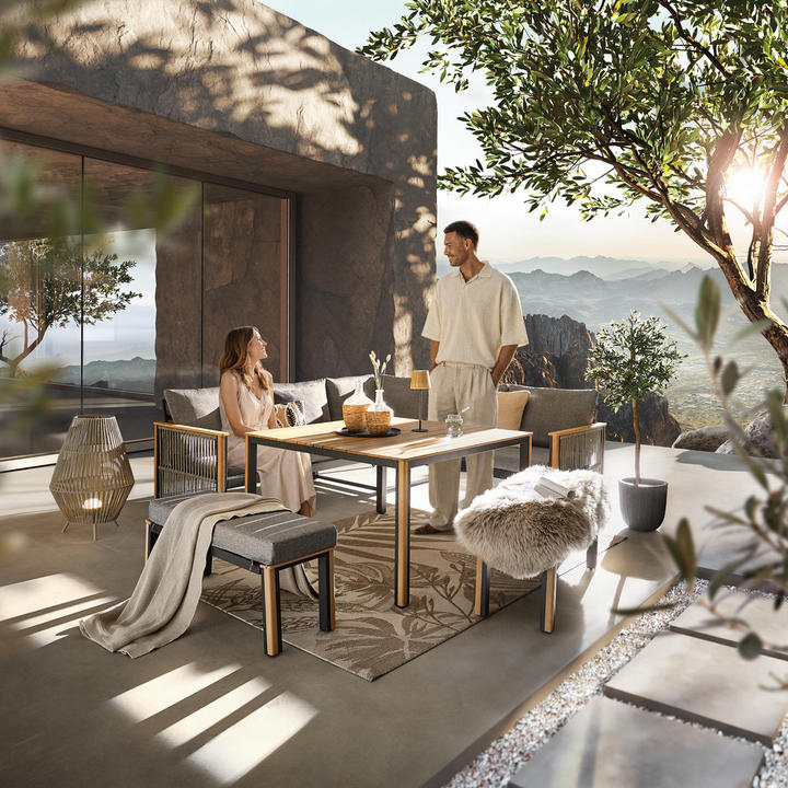 Zwei Personen sitzen und stehen an einem modernen Gartentisch auf einer Terrasse mit Blick auf Berge und Pflanzen.