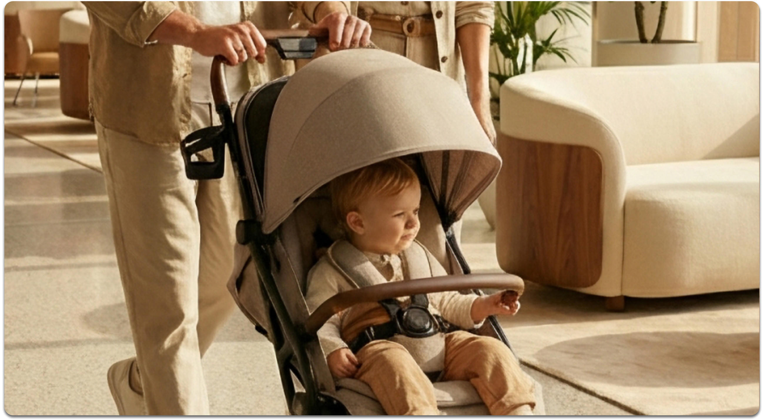 Ein Elternpaar schiebt ihr Kind im Kinderwagen einen Flur entlang. Der Boden besteht aus beigen Fliesen und seitlich stehen Pflanzen und Sessel vor großen Fenstern.