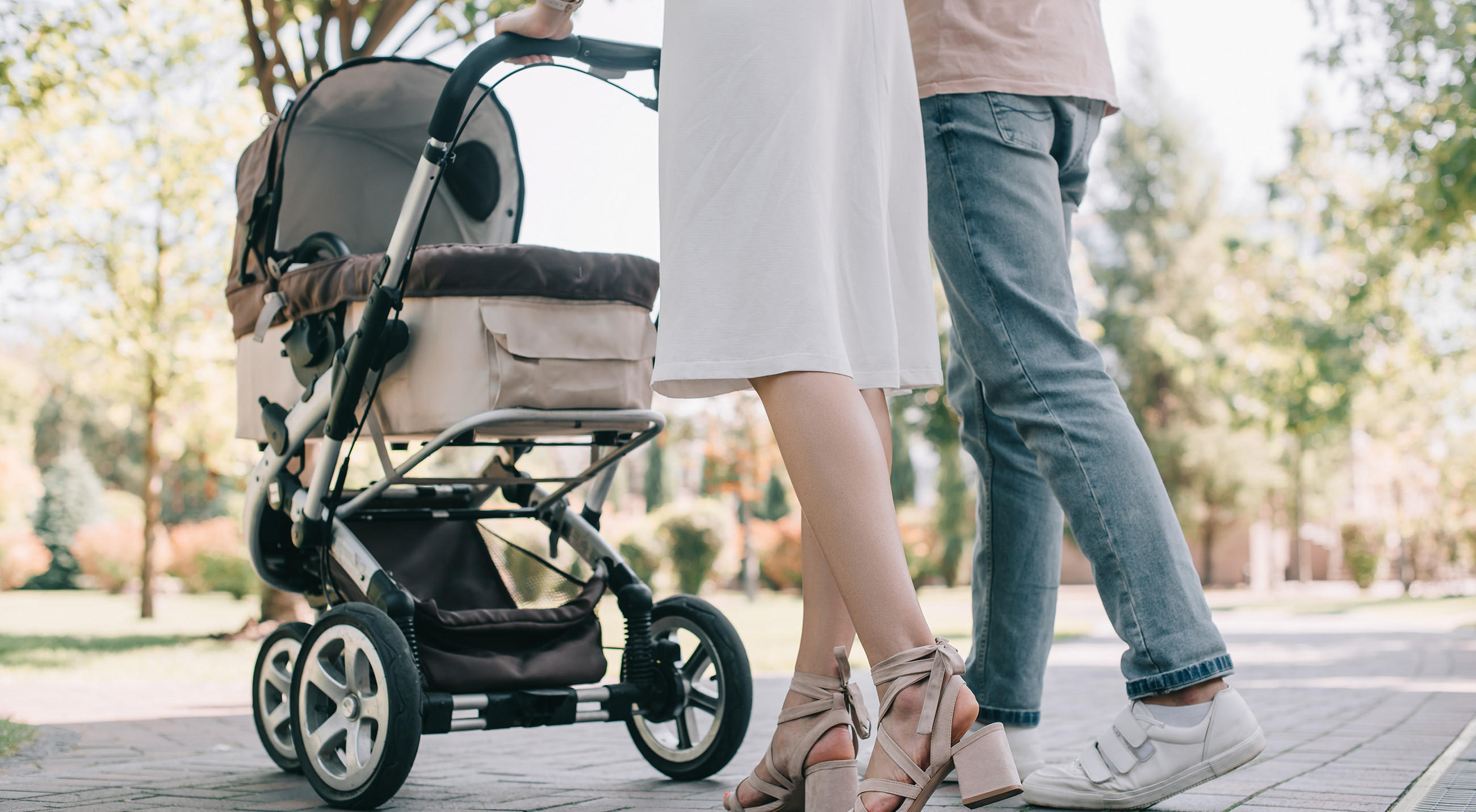 Zwei Personen gehen mit einem Kinderwagen durch einen Park, sichtbar sind ihre Beine und Schuhe sowie die großen Räder des Outdoor-Kinderwagens