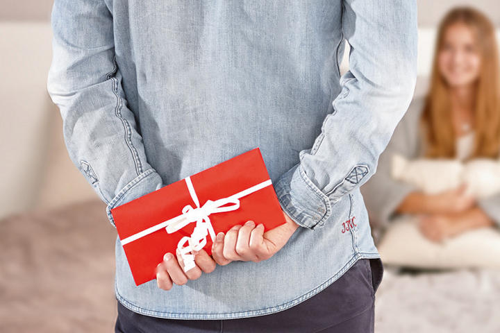 Person mit rotem Geschenkumschlag hinter dem Rücken, daneben vier farbige XXXL-Gutscheinkarten – ideal für stilvolle Geschenkideen rund ums Wohnen.