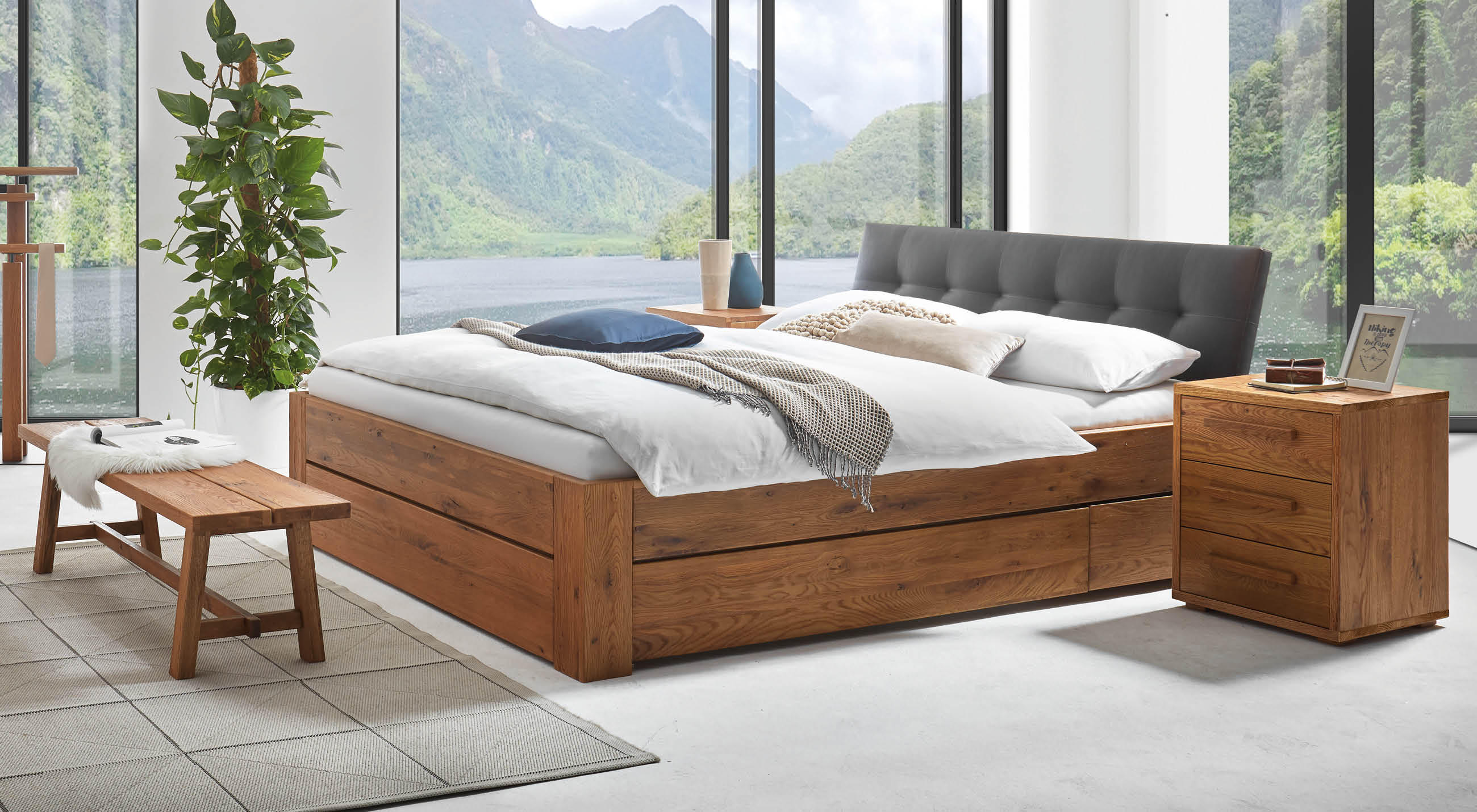 Hasena Stauraum-Bett aus Massivholz mit integrierten Schubladen und gepolstertem Kopfteil, inszeniert in einem hellen Schlafzimmer mit Panorama-Fenstern und Bergsee-Blick.
